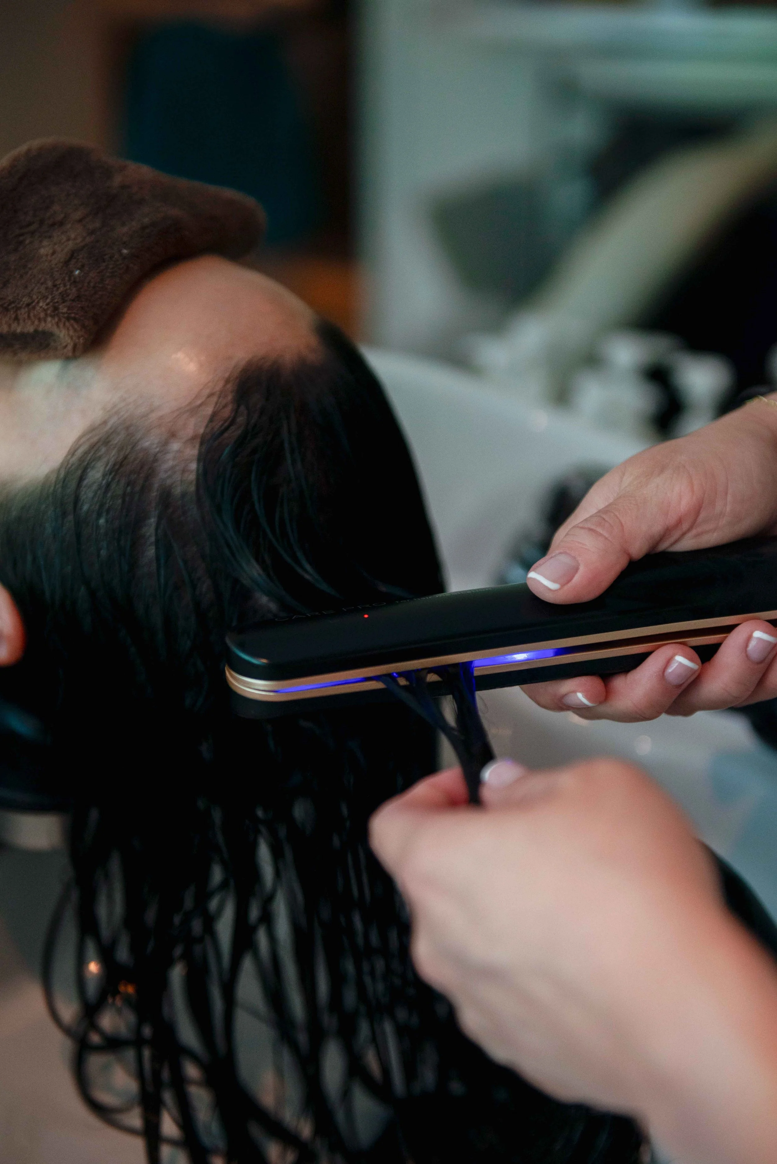 Person getting hair treatment with a heated iron applied to their wet, dark hair in a salon setting.