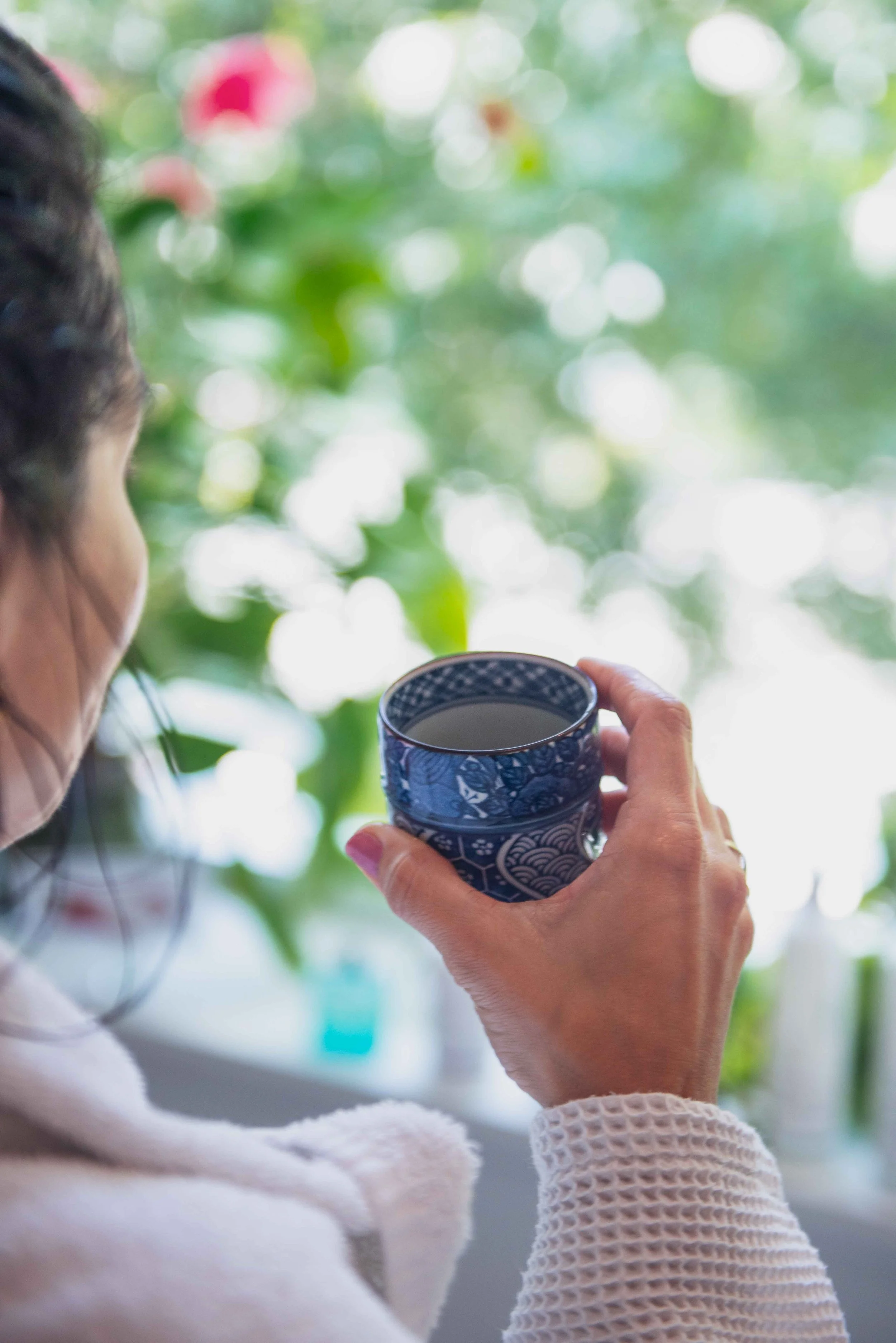 Person holding a tea cup in front of a window with green leaves outside.
