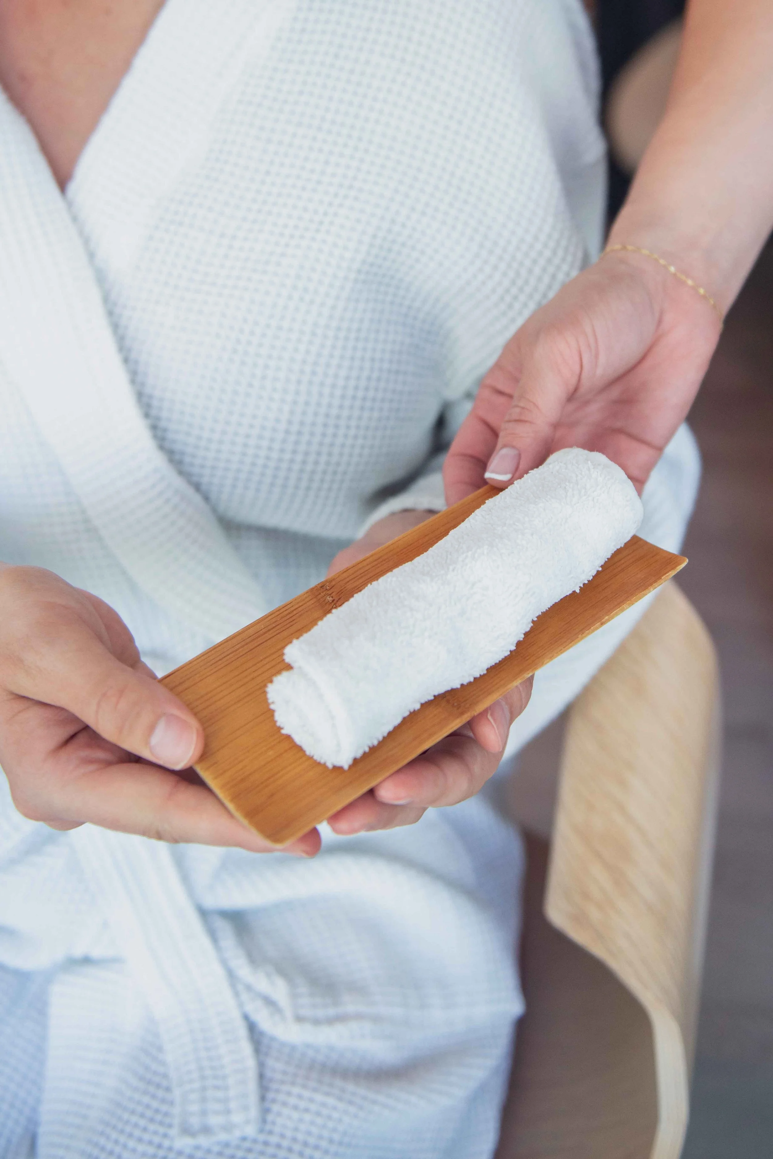 Close-up of a person holding a wooden tray with a rolled white towel, wearing a white textured robe.
