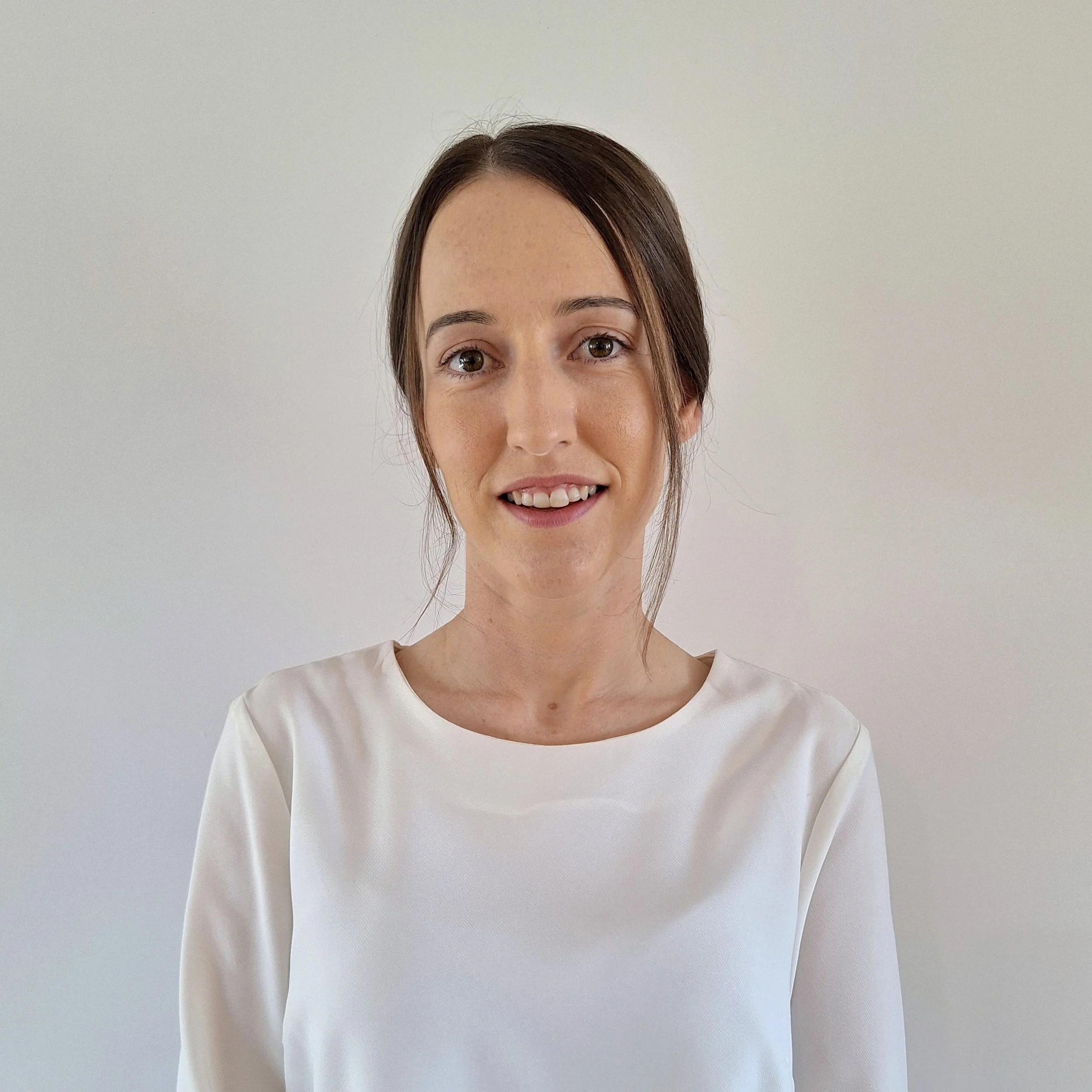 Portrait of a young woman with brown hair in a loose ponytail, wearing a white satin blouse, smiling softly against a plain white wall.