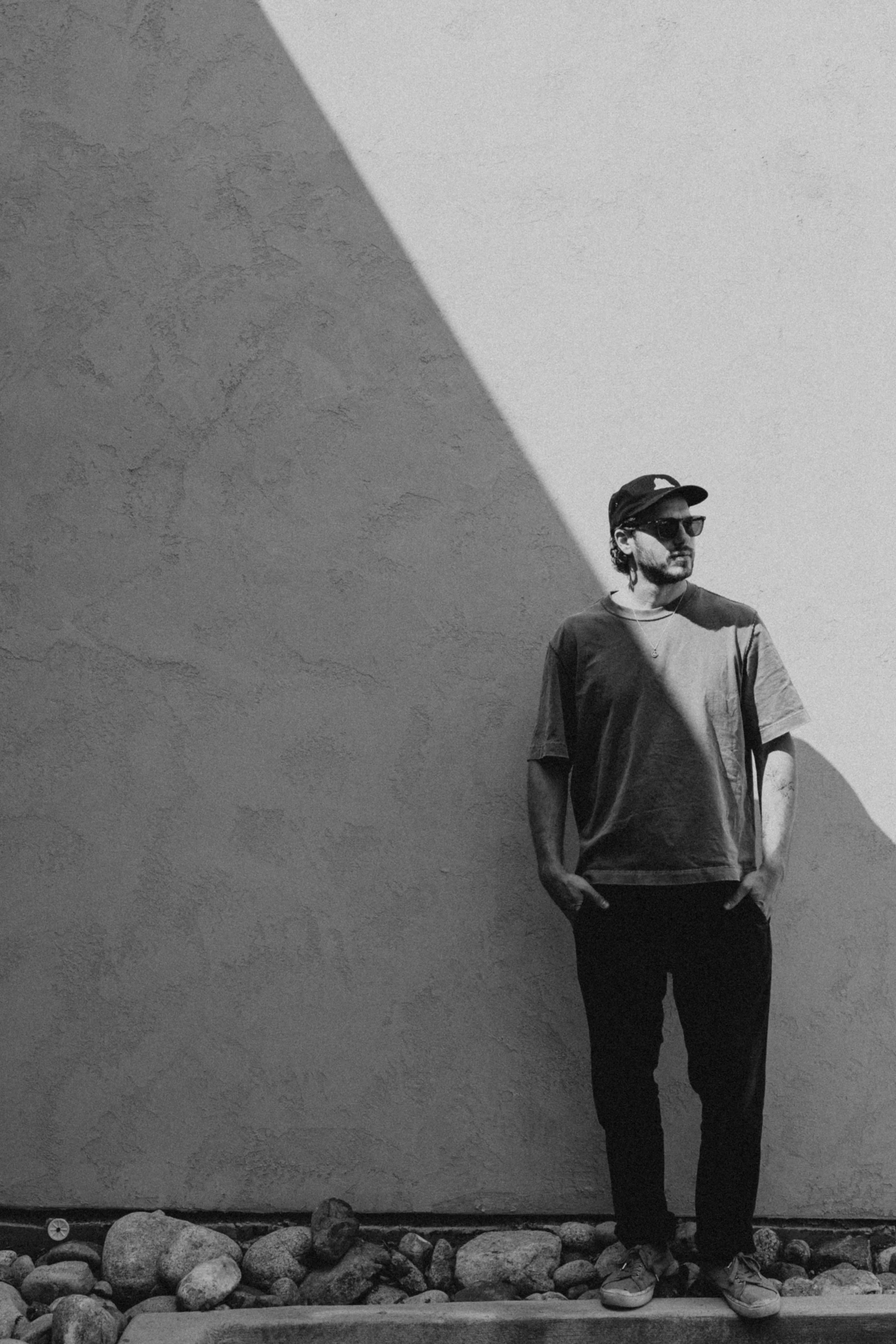 Matt Jalbert wearing sunglasses and a cap stands against a wall with a shadow line at an angle, with rocks at his feet.