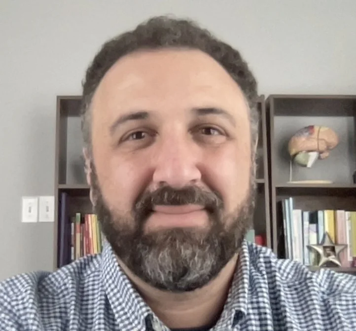 Man with a beard and checked shirt, bookshelves with books and a brain model in the background.