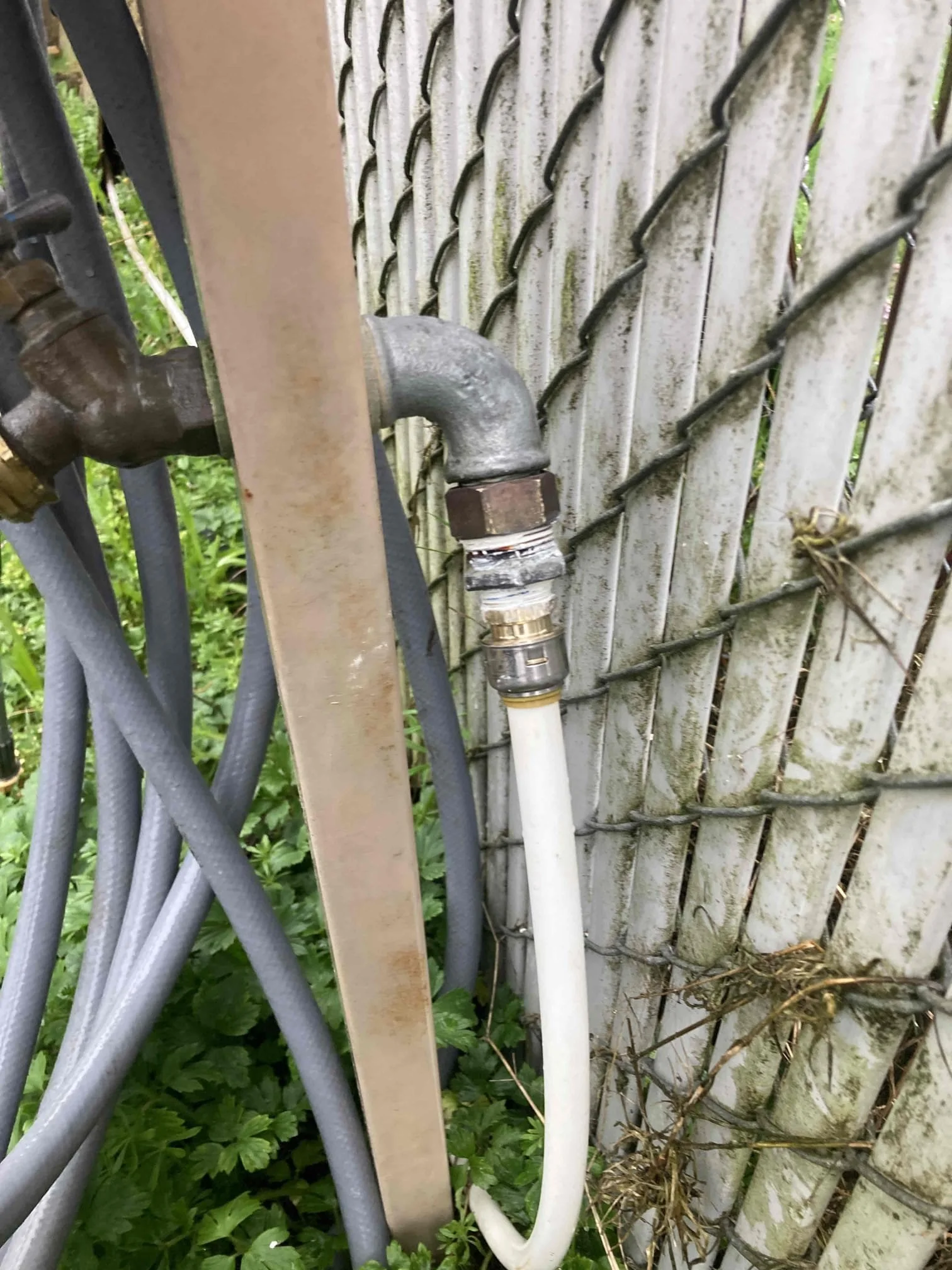 Close-up of a water spigot and hose connected to an outdoor faucet, with a chain-link fence and greenery in the background.
