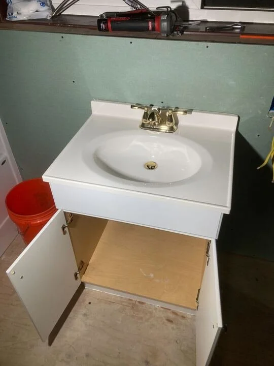 A white bathroom vanity with a sink and faucet, open cabinet door revealing an empty shelf, in a room with a green wall and various tools on the countertop.