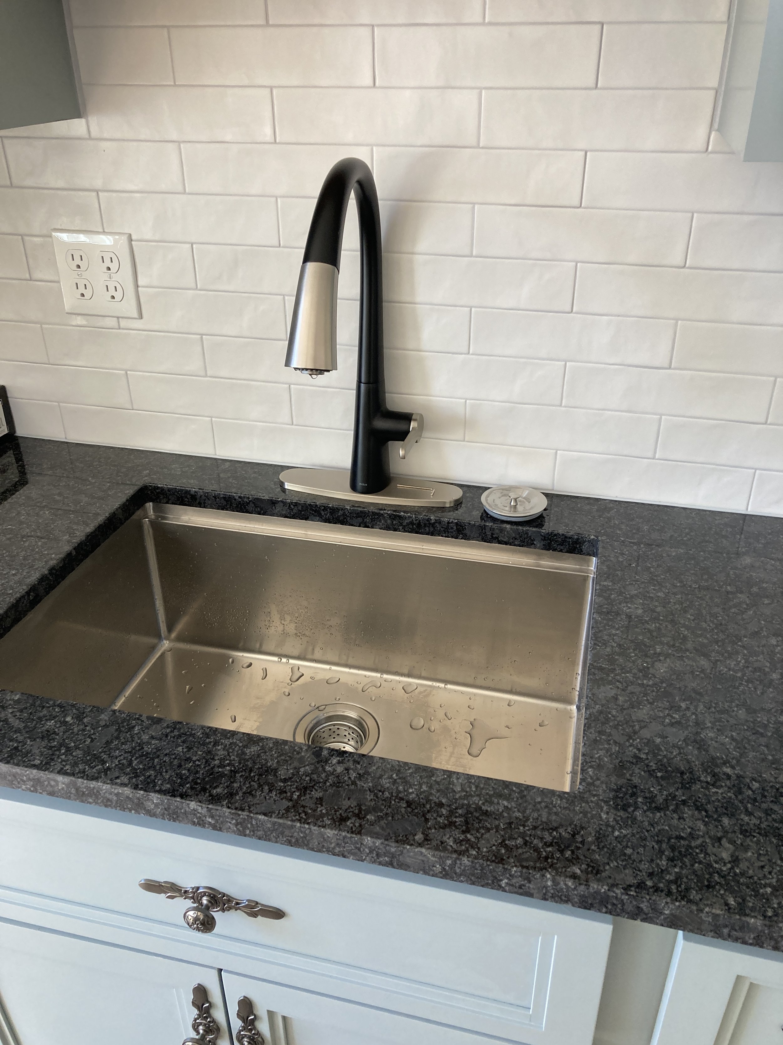 A kitchen sink with a black and silver faucet, set into a black granite countertop, with a white tiled backsplash and white cabinets below.