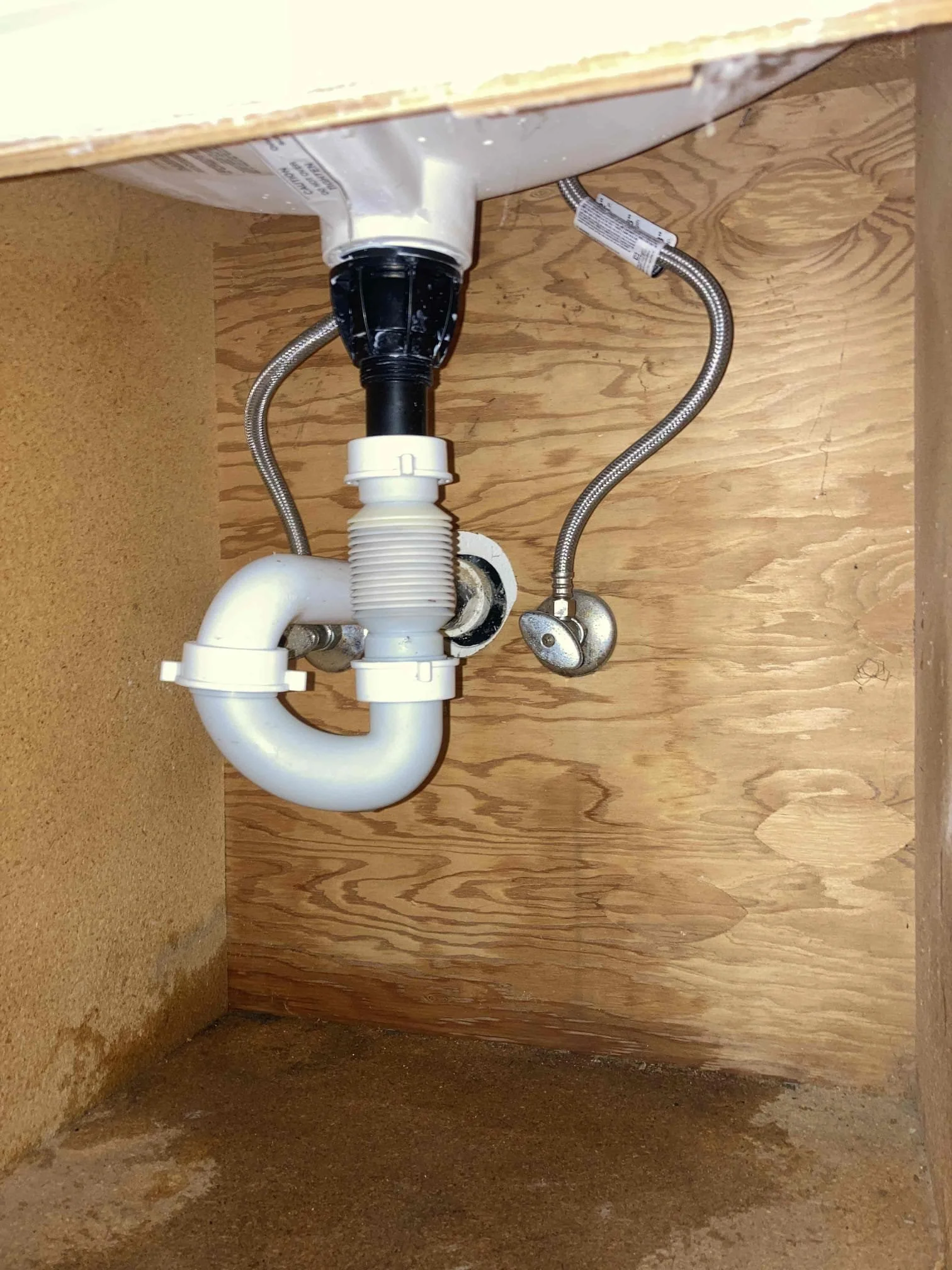 Underneath a kitchen sink showing plumbing pipes, including a white P-trap, drain pipe, supply lines, and part of the sink basin.