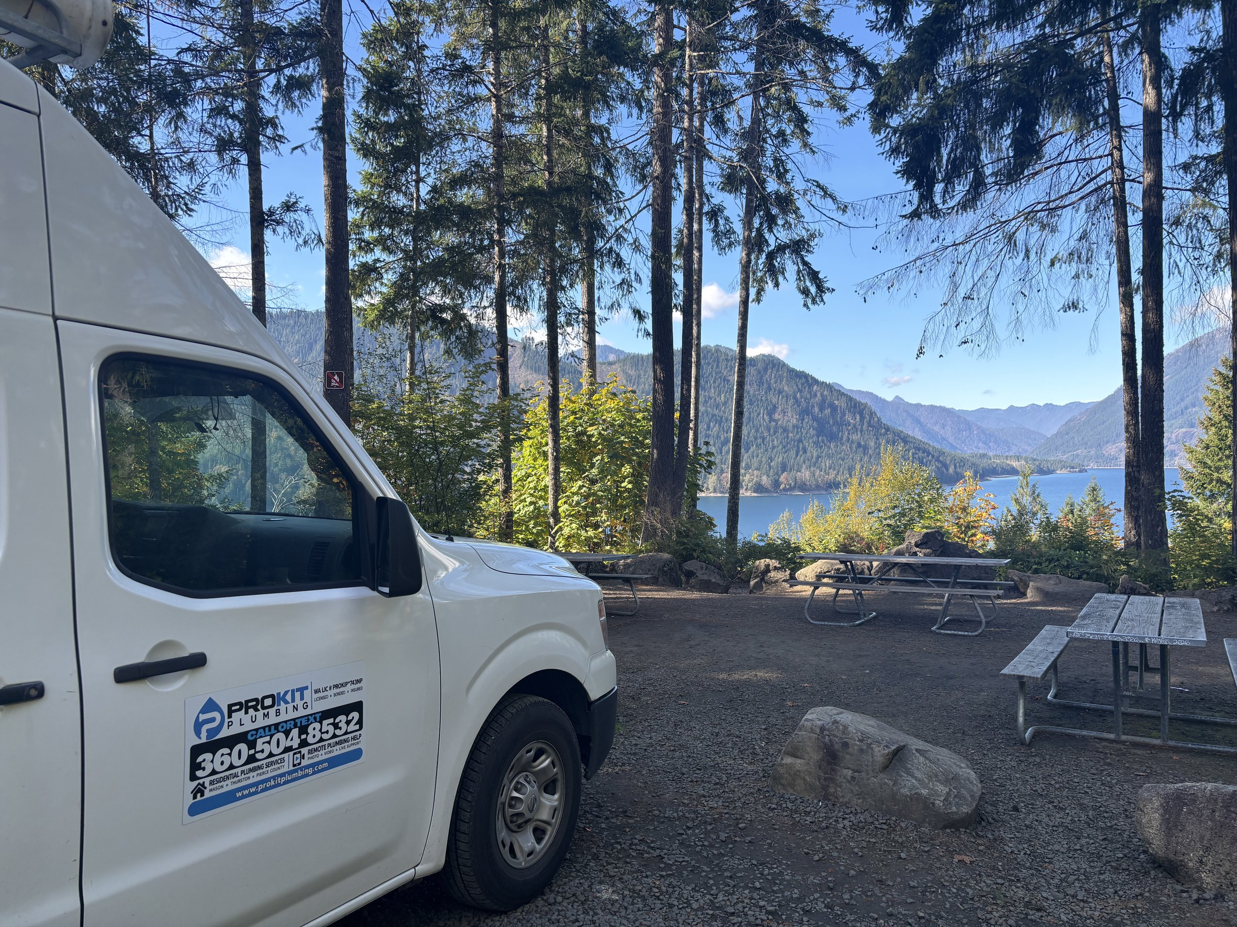 Part of a white ProKit Plumbing service van with contact info, parked in a forested area with trees, mountains, and a lake in the background.