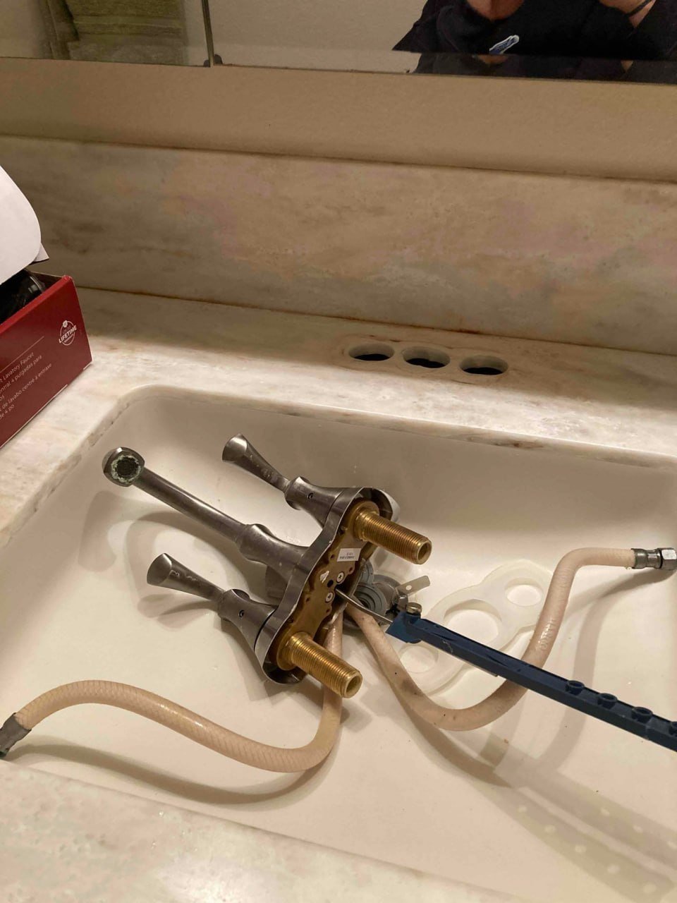 Disassembled faucet parts with hoses placed in a bathroom sink with a mirror above, reflecting part of a person's head and upper body.