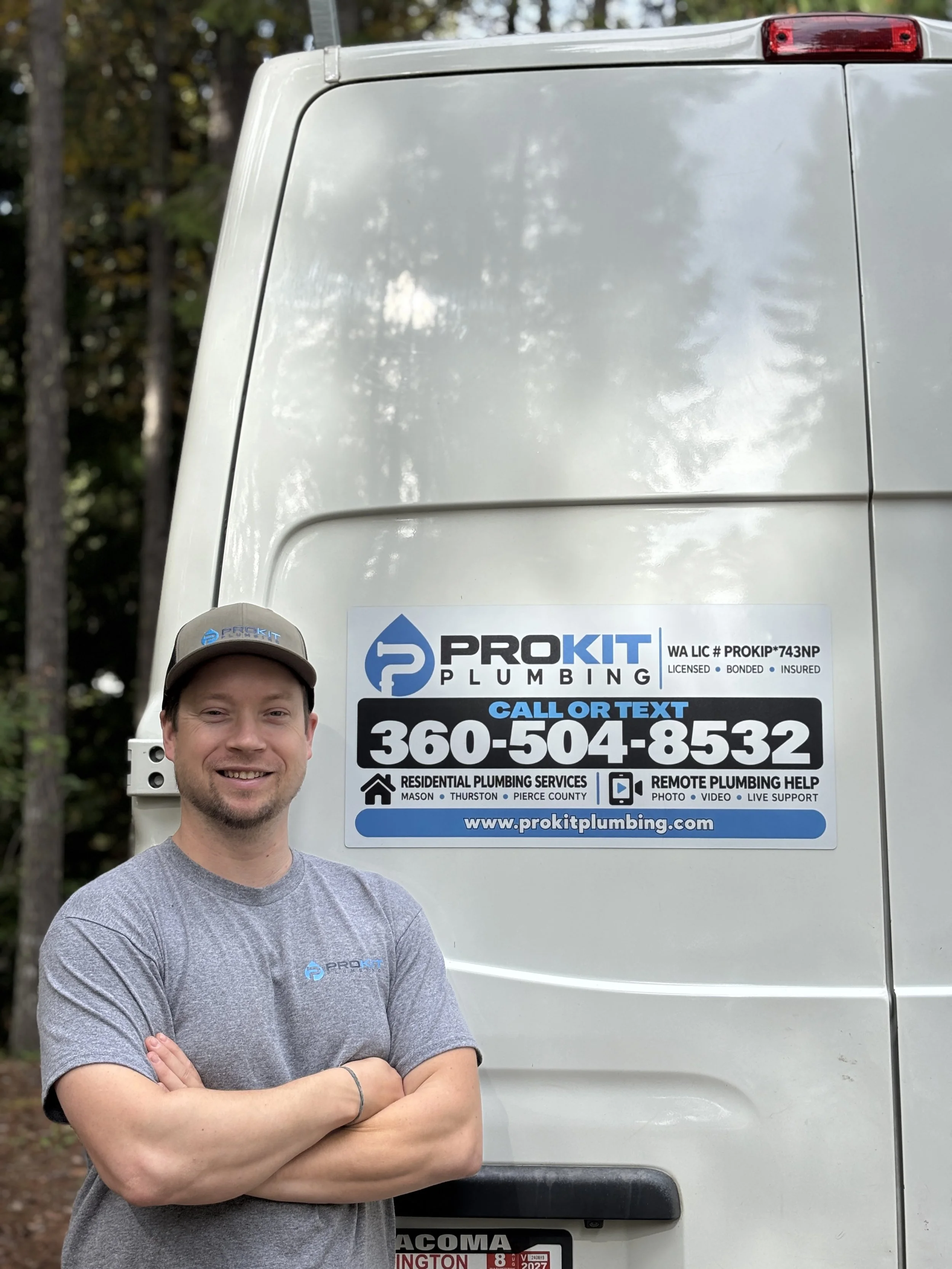 A man with crossed arms standing in front of a white ProKit Plumbing van, smiling, outdoors with trees in the background.