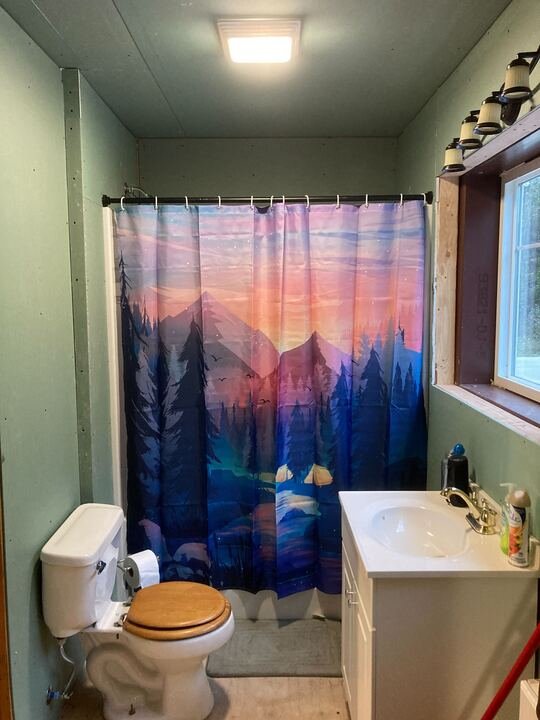 Small bathroom with a mountain landscape shower curtain featuring mountains and trees during a colorful sunset. Includes a toilet with a wooden seat, a white sink with toiletries, a window, and a light fixture.