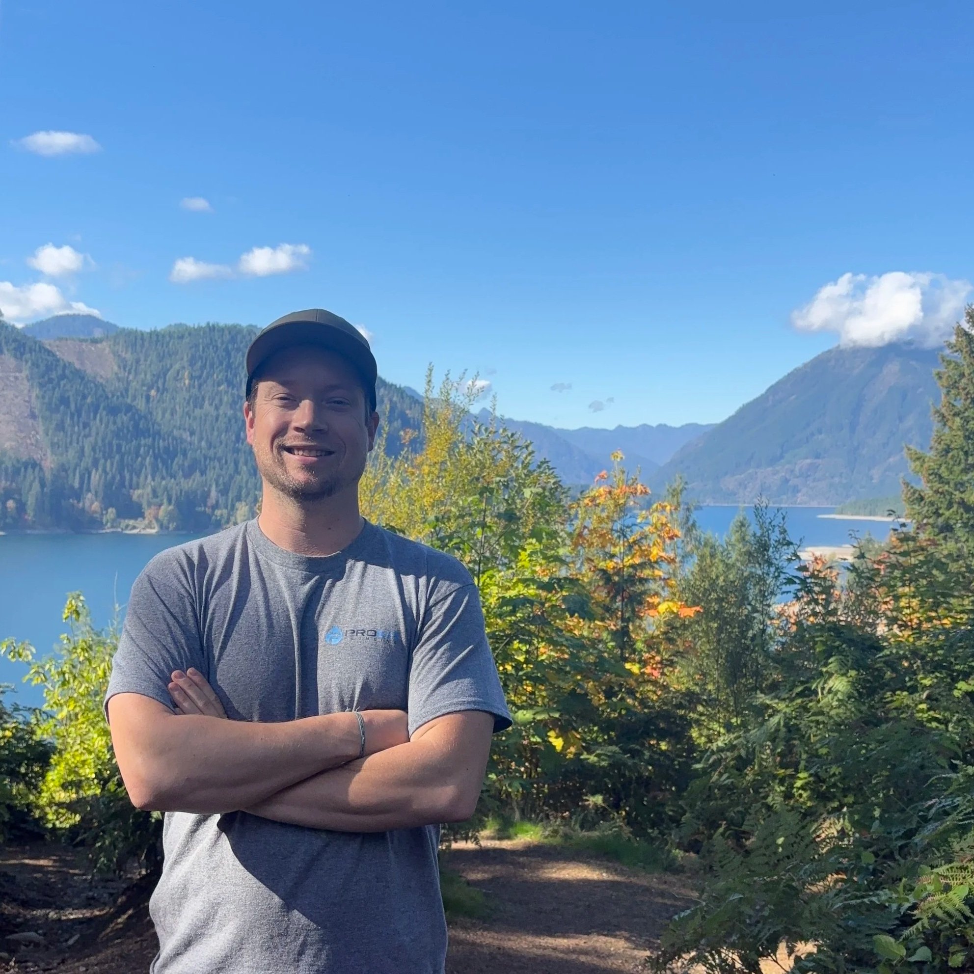 Smiling man in gray t-shirt and baseball cap standing outdoors with arms crossed, trees, mountains, and blue sky with clouds in the background.