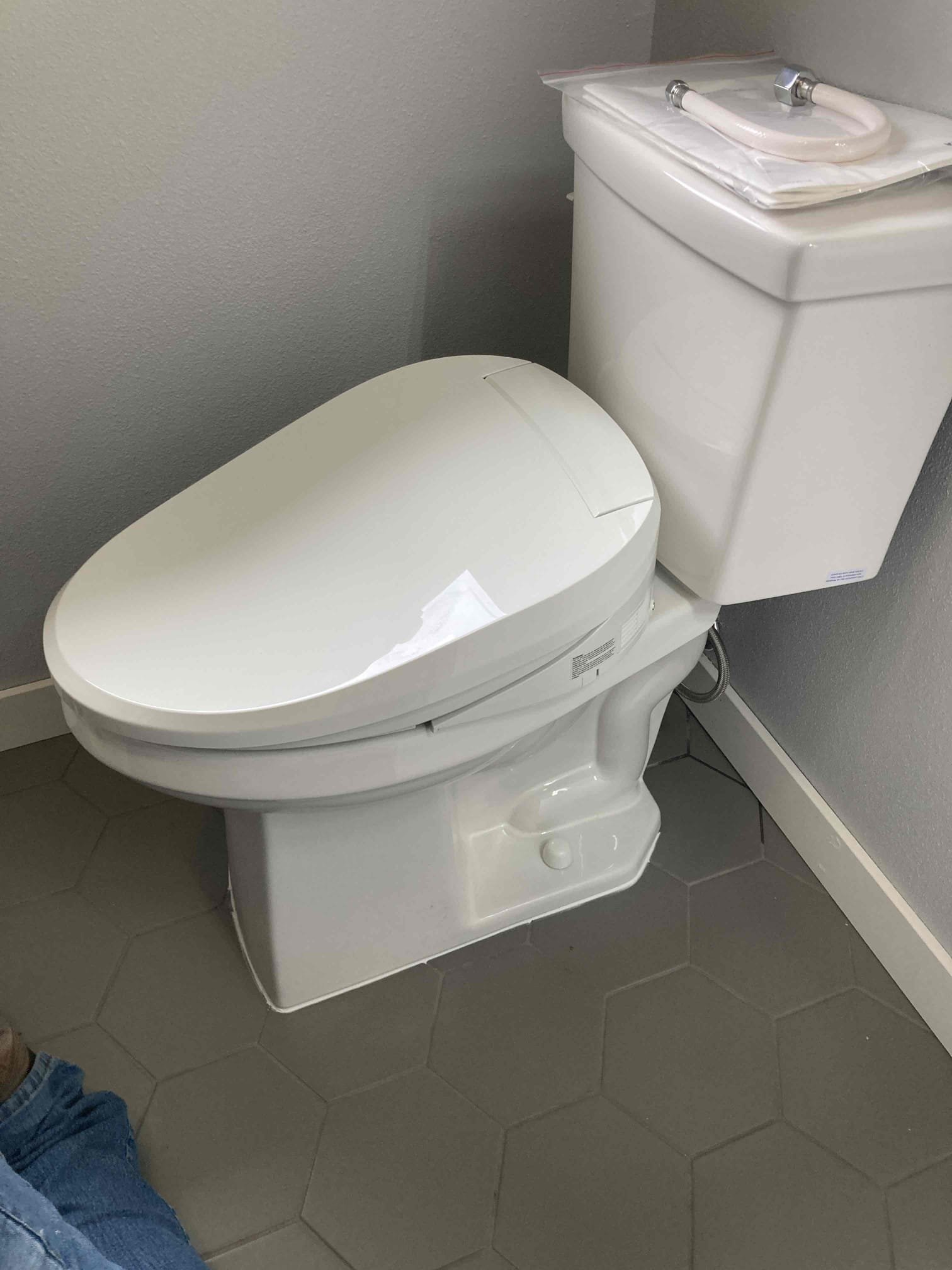 A modern white toilet with a bidet lid, positioned against a gray wall, with hexagonal gray floor tiles.