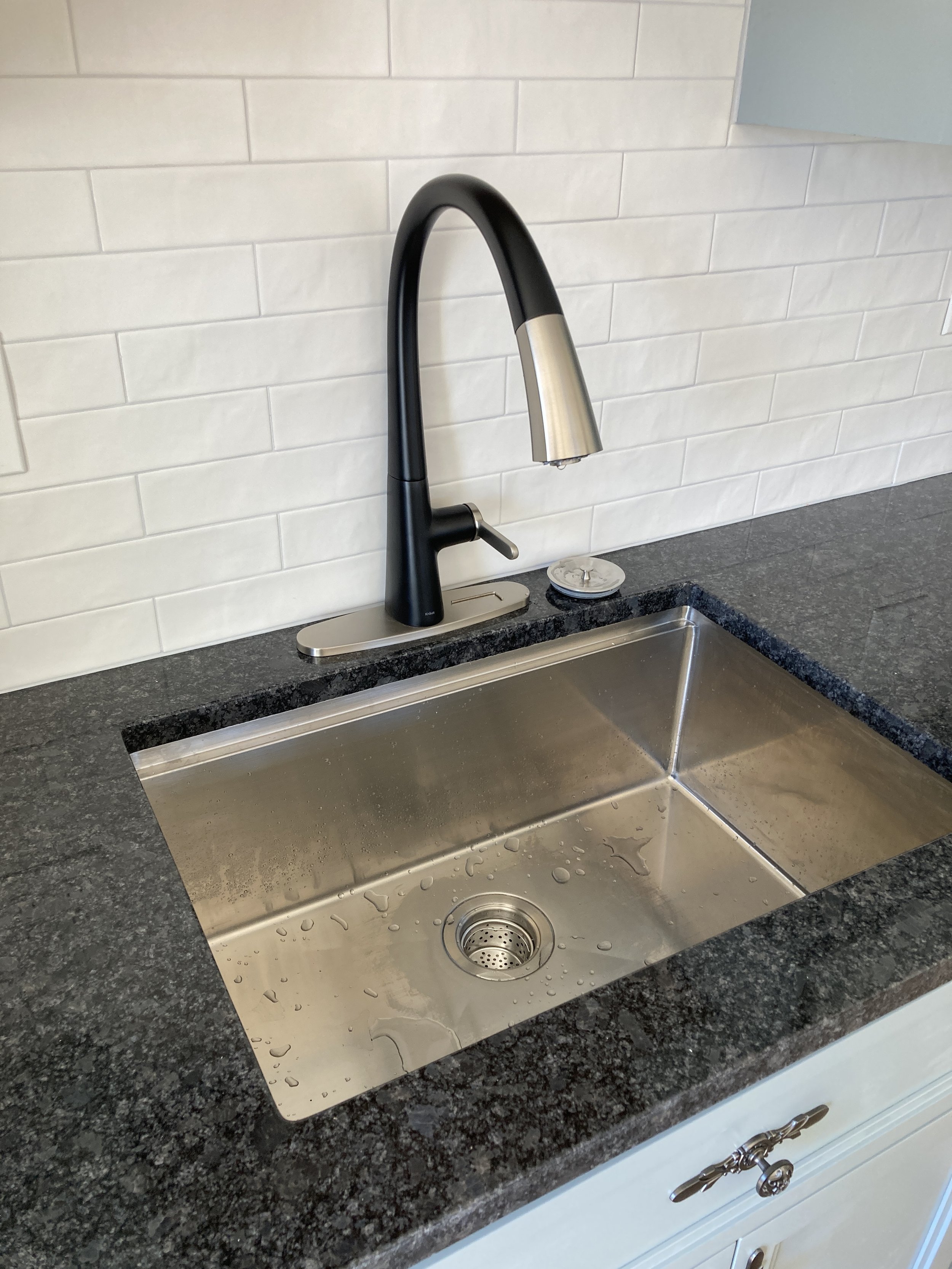 A kitchen stainless steel sink with a black and silver gooseneck faucet and a soap dispenser on a black granite countertop.