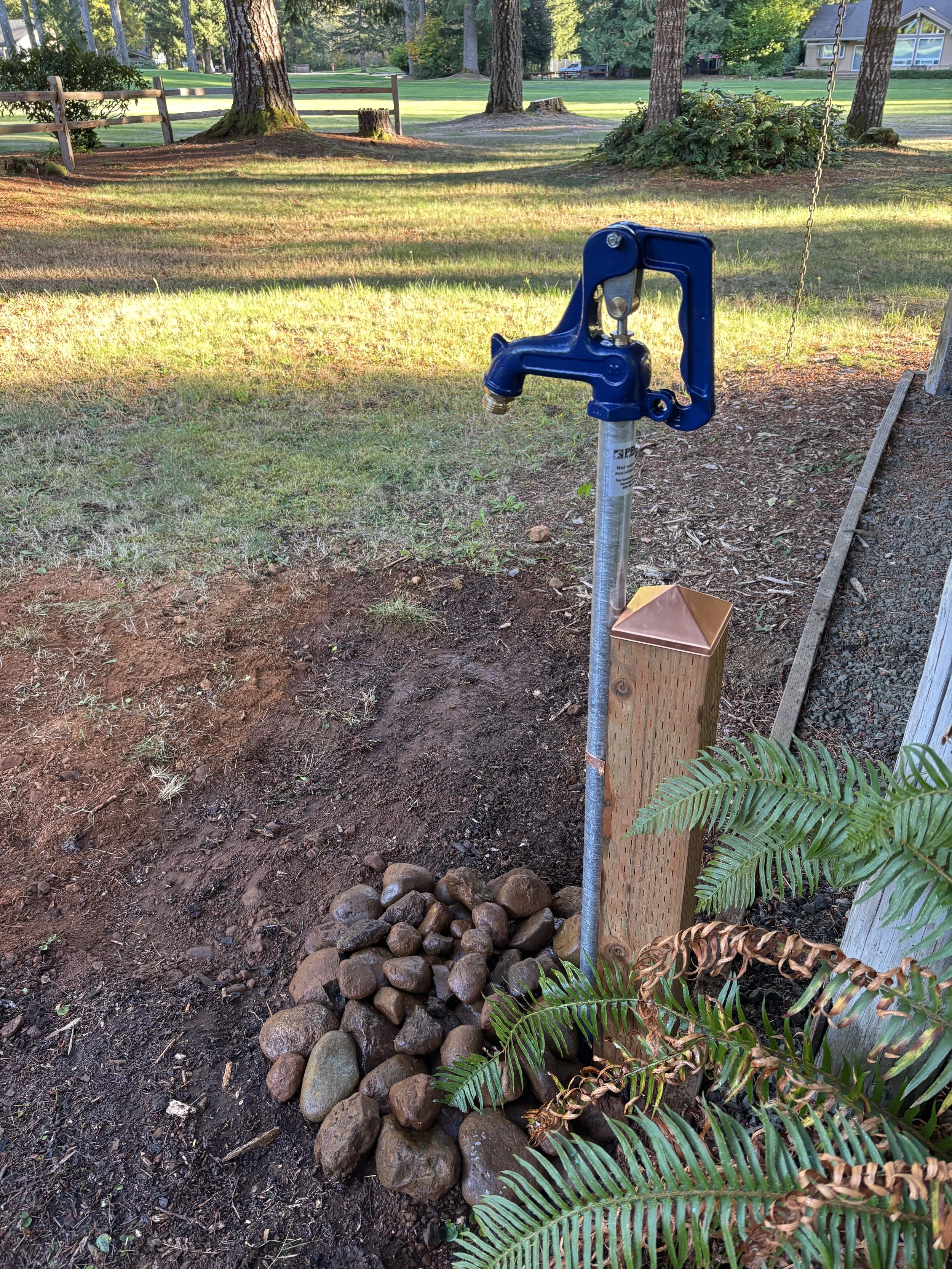 A blue water spigot on a metal post with a string attached, situated on a landscaped area with a pile of large rocks nearby, surrounded by plants and trees in a park or backyard.