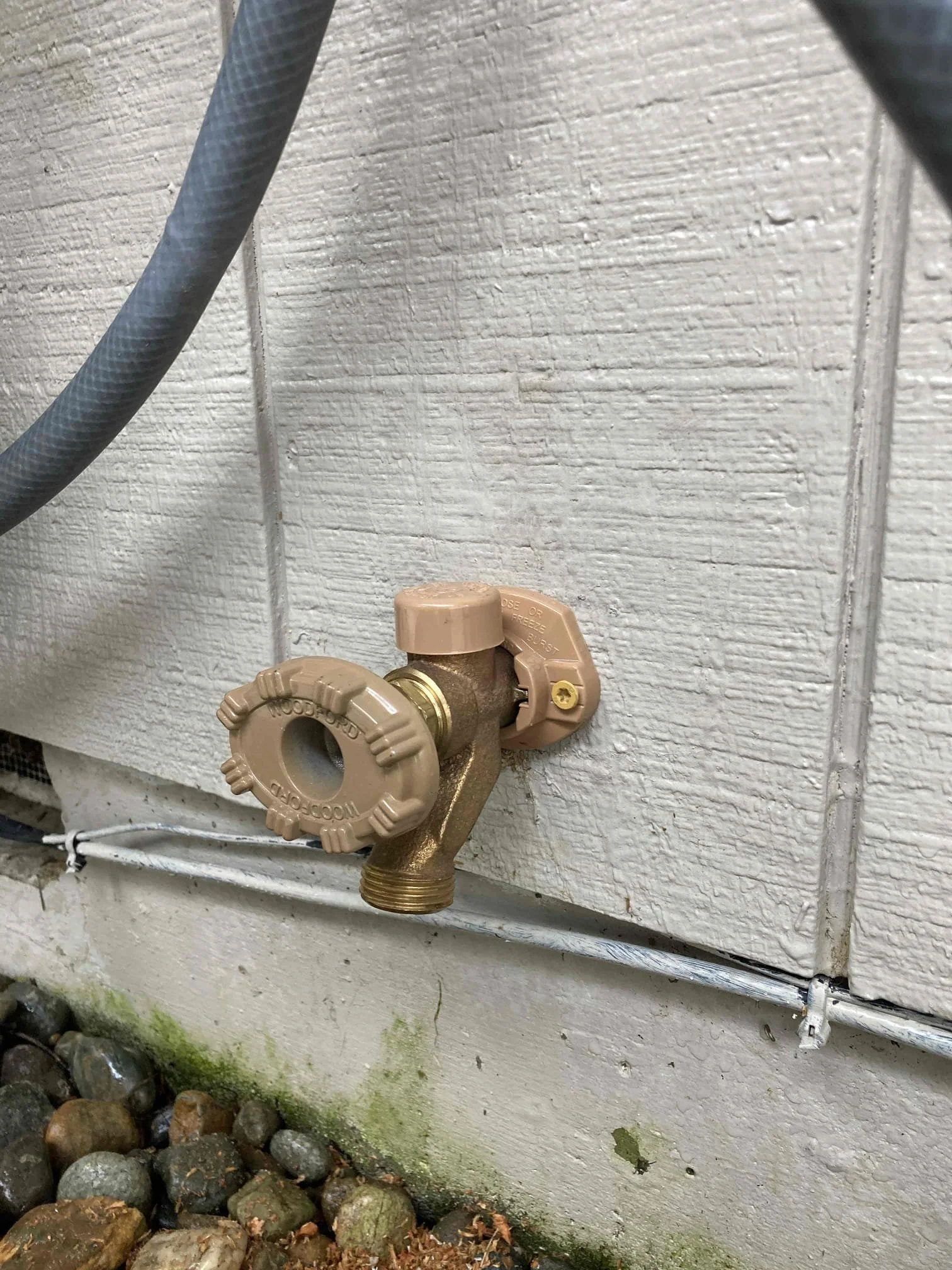 Outdoor water faucet with beige handle attached to a white wooden wall, near gray electrical conduit and rocky ground with moss patches.