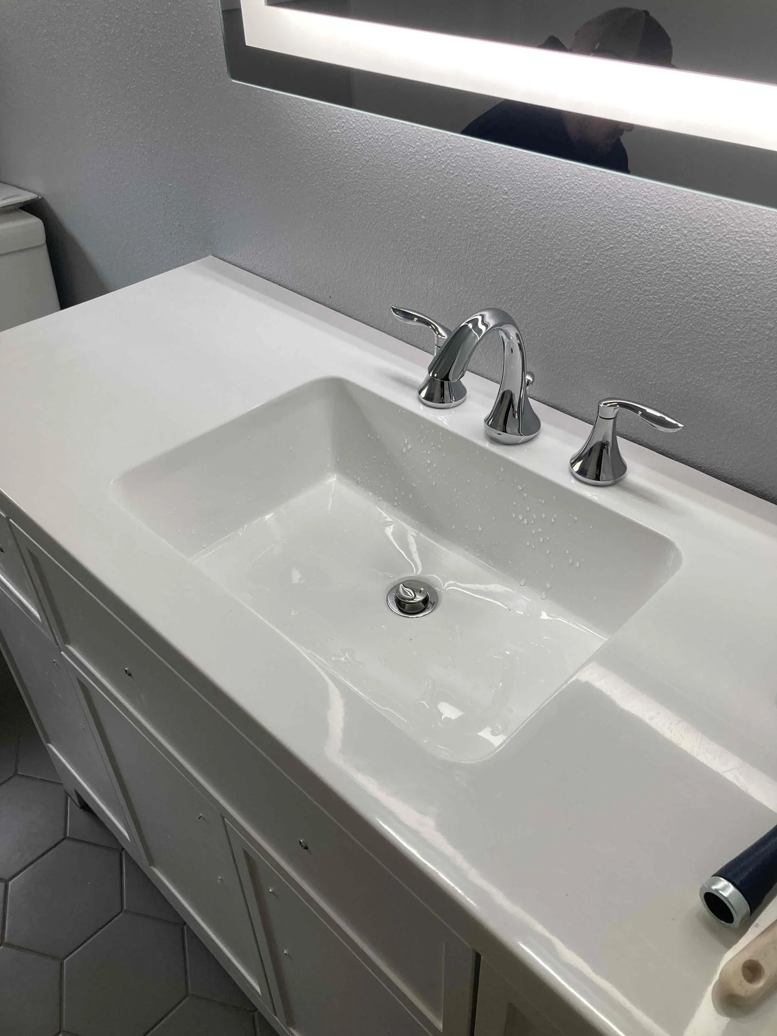 Bathroom sink with chrome faucet, mounted on white vanity with cabinet doors, and gray hexagonal tiled floor.