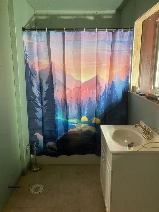 Bathroom with green walls, a shower curtain depicting a mountain and forest scene at sunset, a white sink with gold fixtures, and a small window on the right.