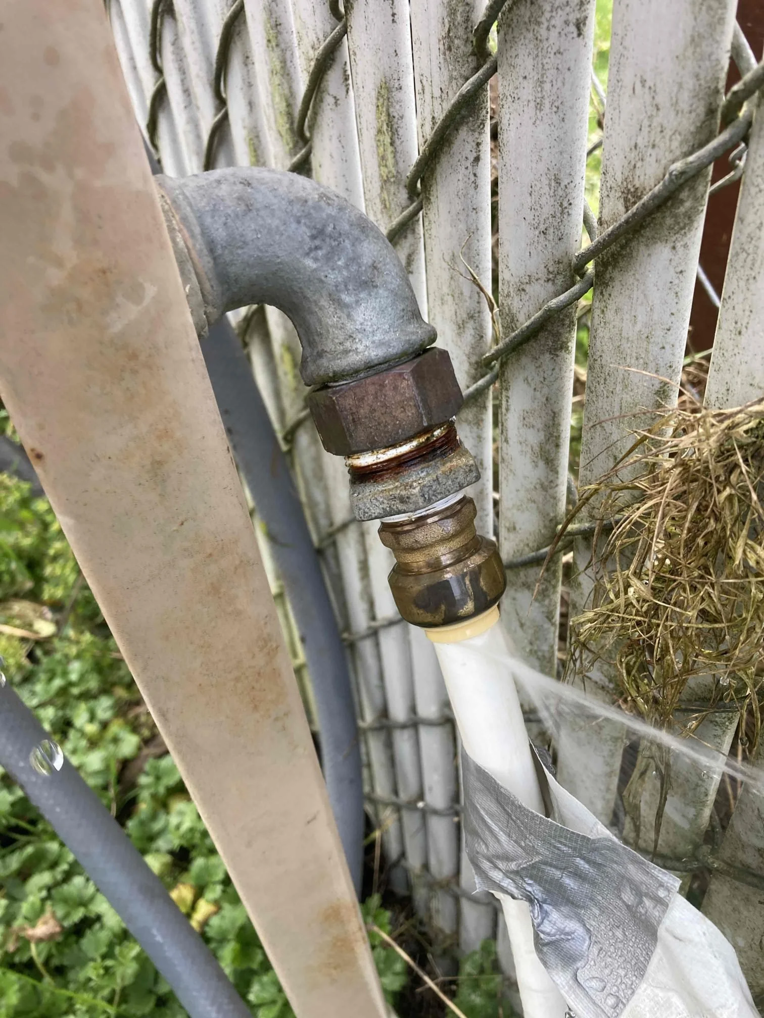 Close-up of a rusty outdoor water pipe connection with a white plastic pipe, attached to a metal fence with green grass and weeds nearby.