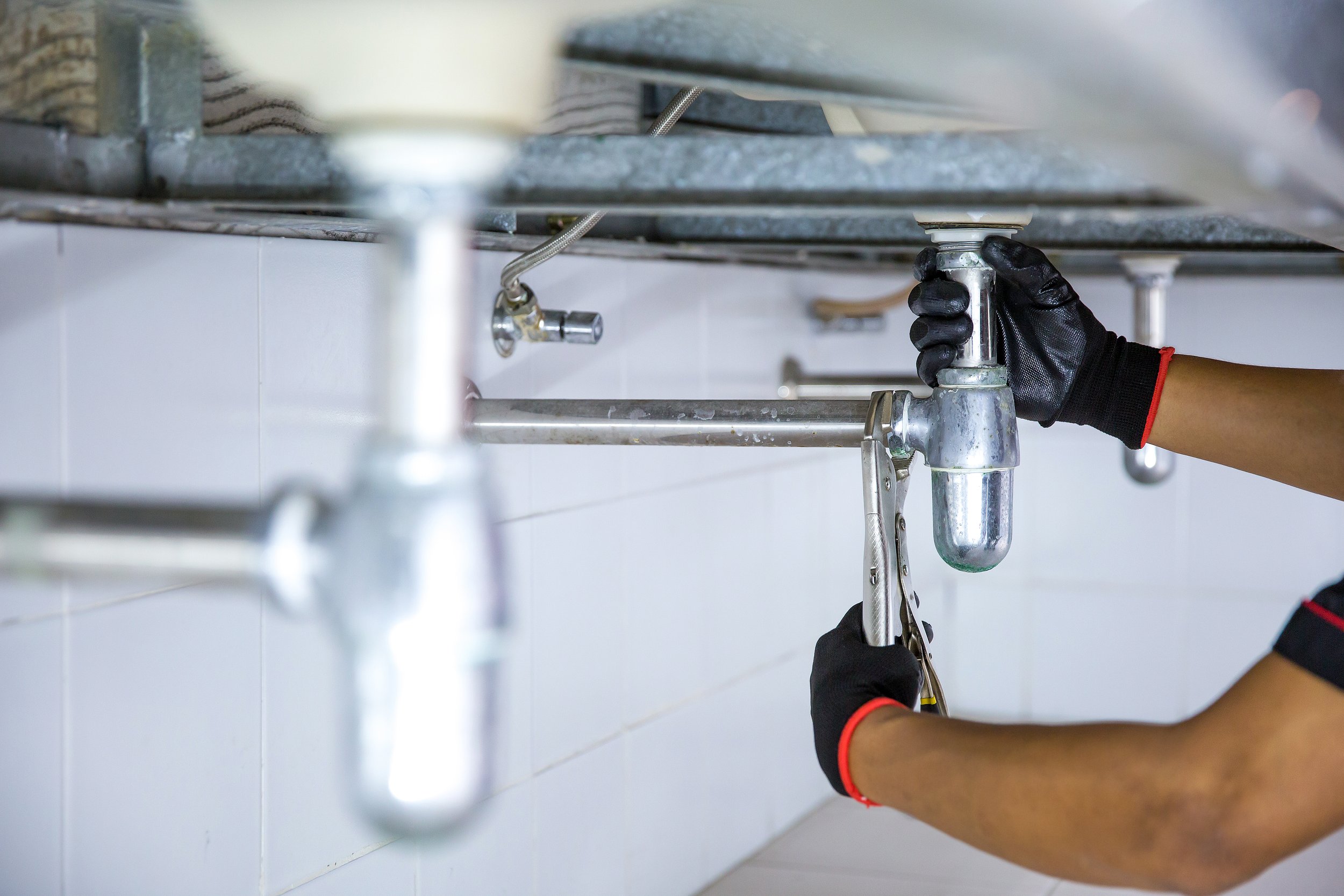 A person wearing black gloves tightening a plumbing pipe under a sink with a wrench.