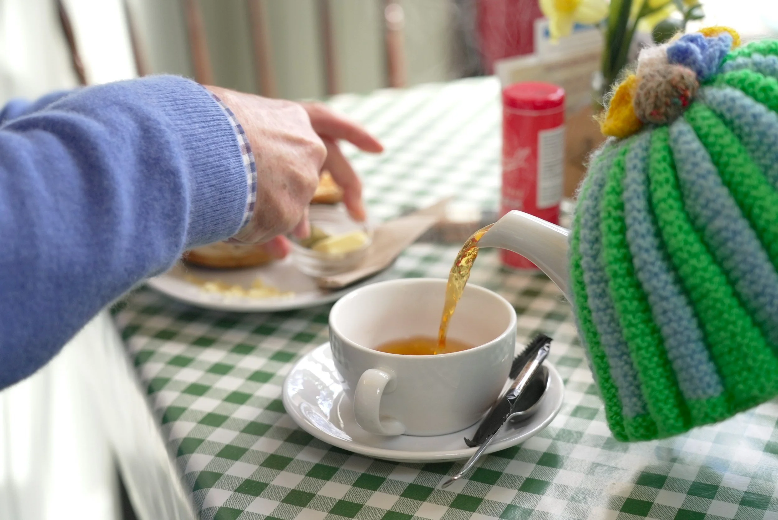 A person pouring tea from a kettle into a white mug on a green and white checkered tablecloth, with a colorful knitted hoodie nearby and breakfast items in the background.