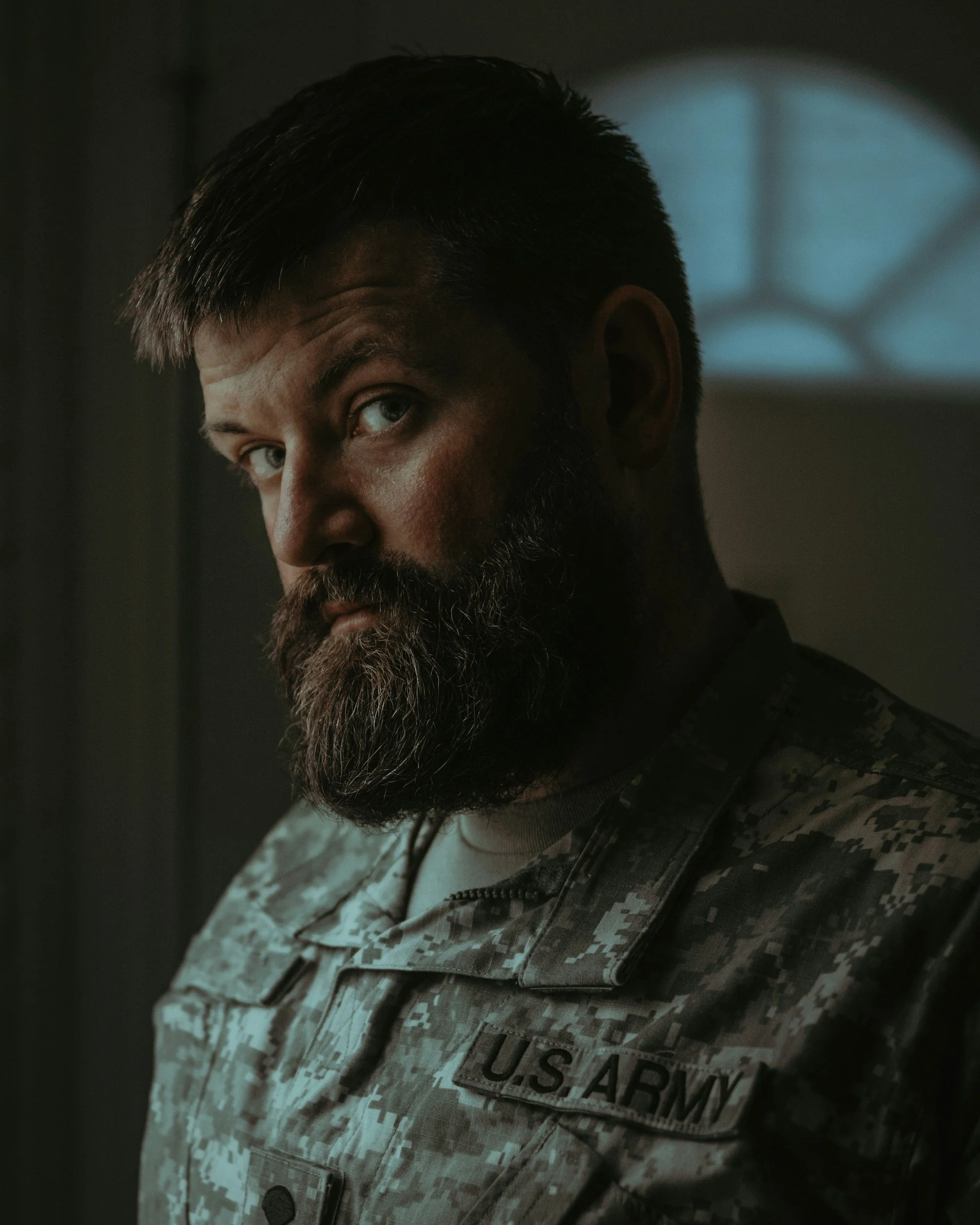 Close-up of a person in military camouflage uniform with a U.S. Army patch on the chest, dark hair, and a beard, in a dimly lit setting.