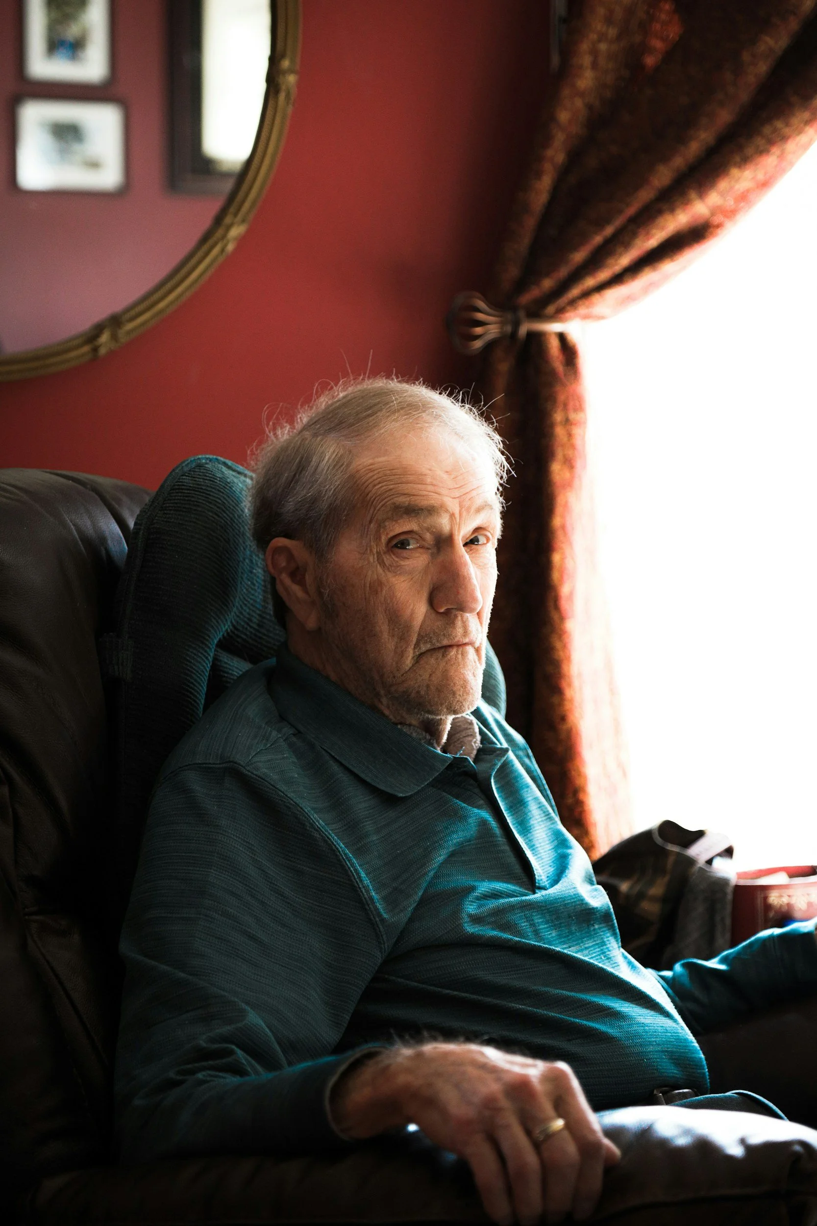 An elderly man with gray hair sitting on a dark brown leather chair near a window with a curtain, looking at the camera with a neutral expression, holding a remote or phone in his right hand, in a room with red walls and framed pictures, sunlight coming through the window.