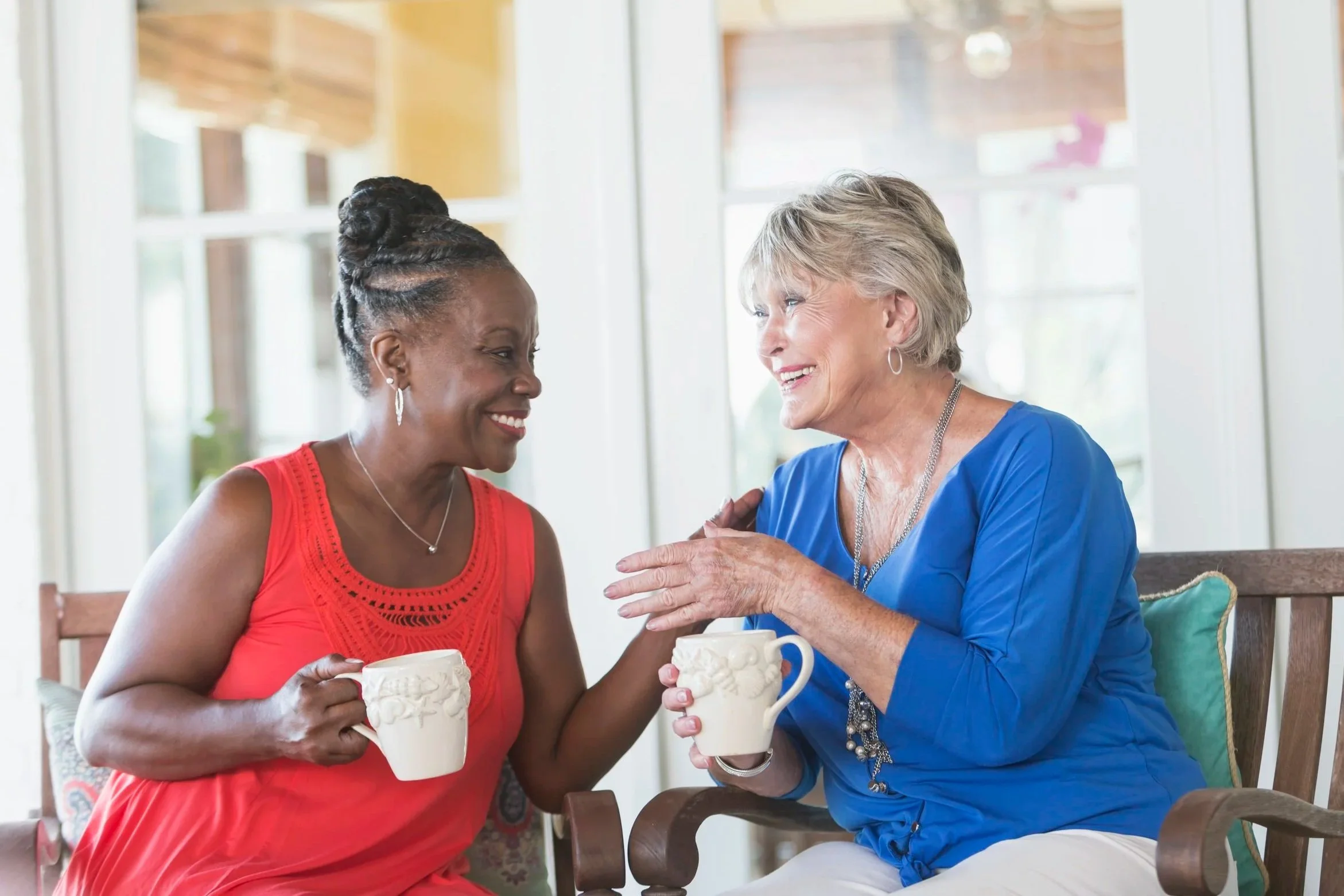 Two women smiling and talking while holding mugs, sitting in front of a bright room with large windows.