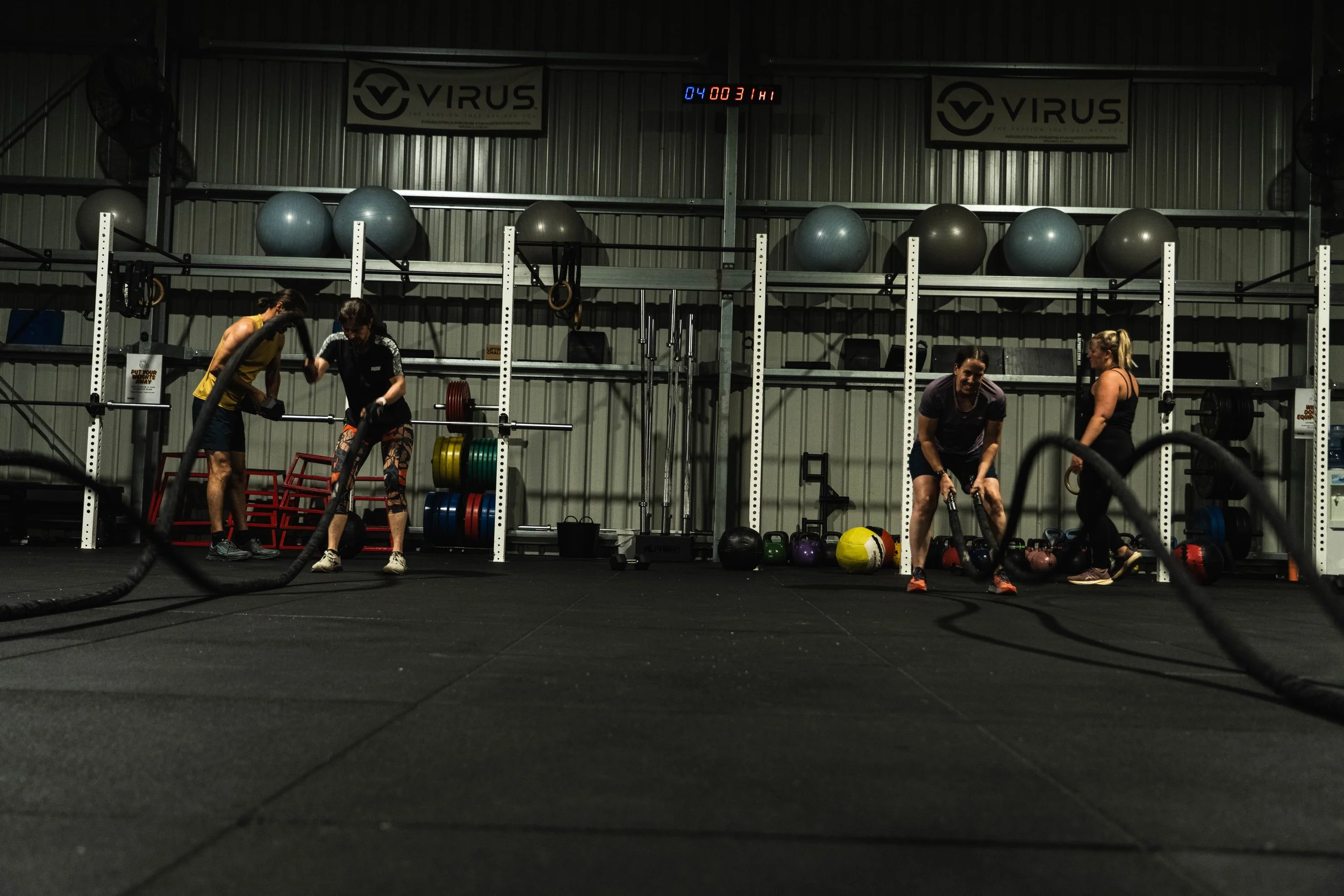 Four women working out with battle ropes in a gym with weight racks and exercise balls in the background.