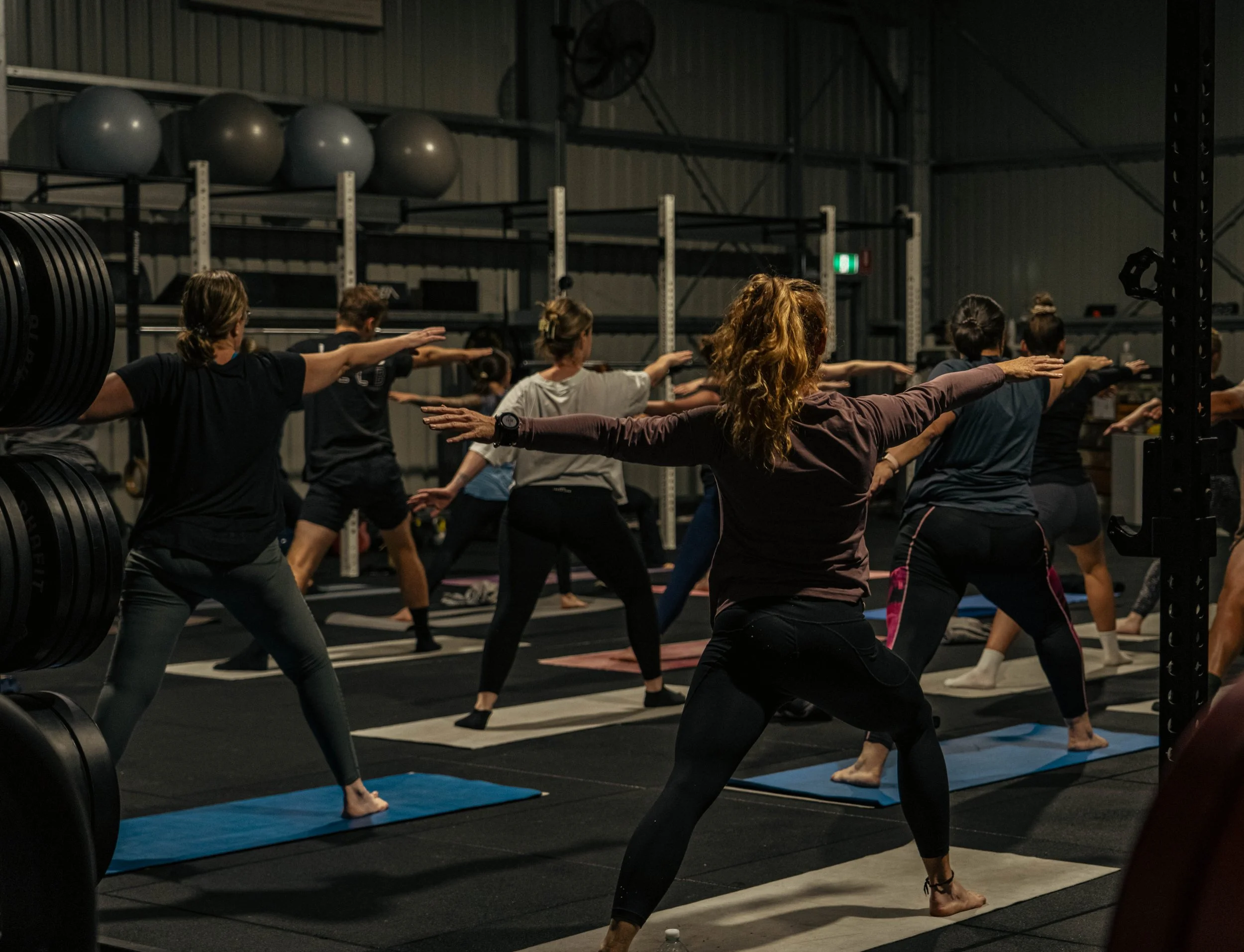 Group of women participating in a yoga or stretching class in a gym with black flooring, mats, and workout equipment, including weights and large exercise balls on shelves.
