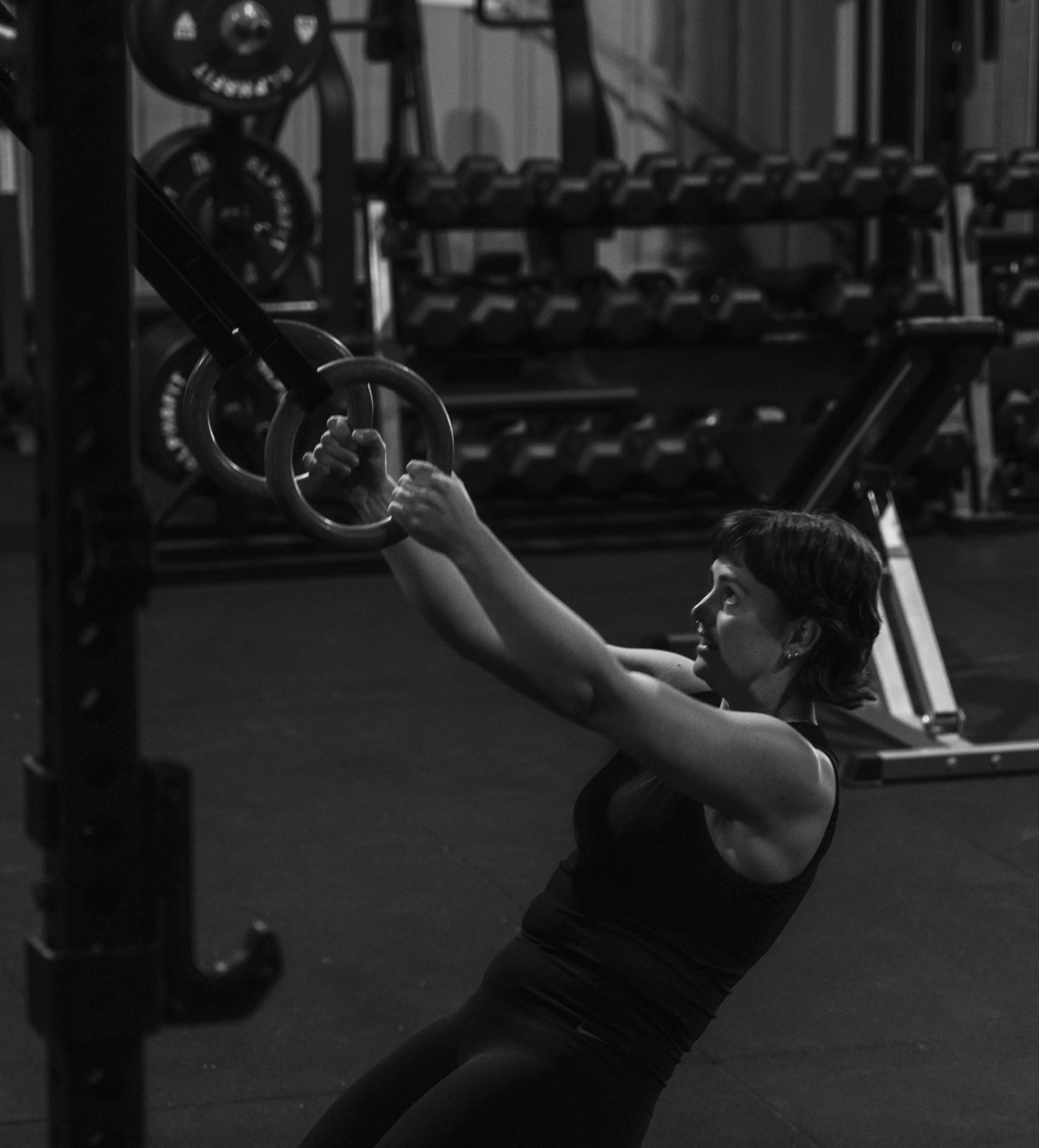 A woman working out at the gym, holding gymnastic rings and lifting herself up, with weights in the background.