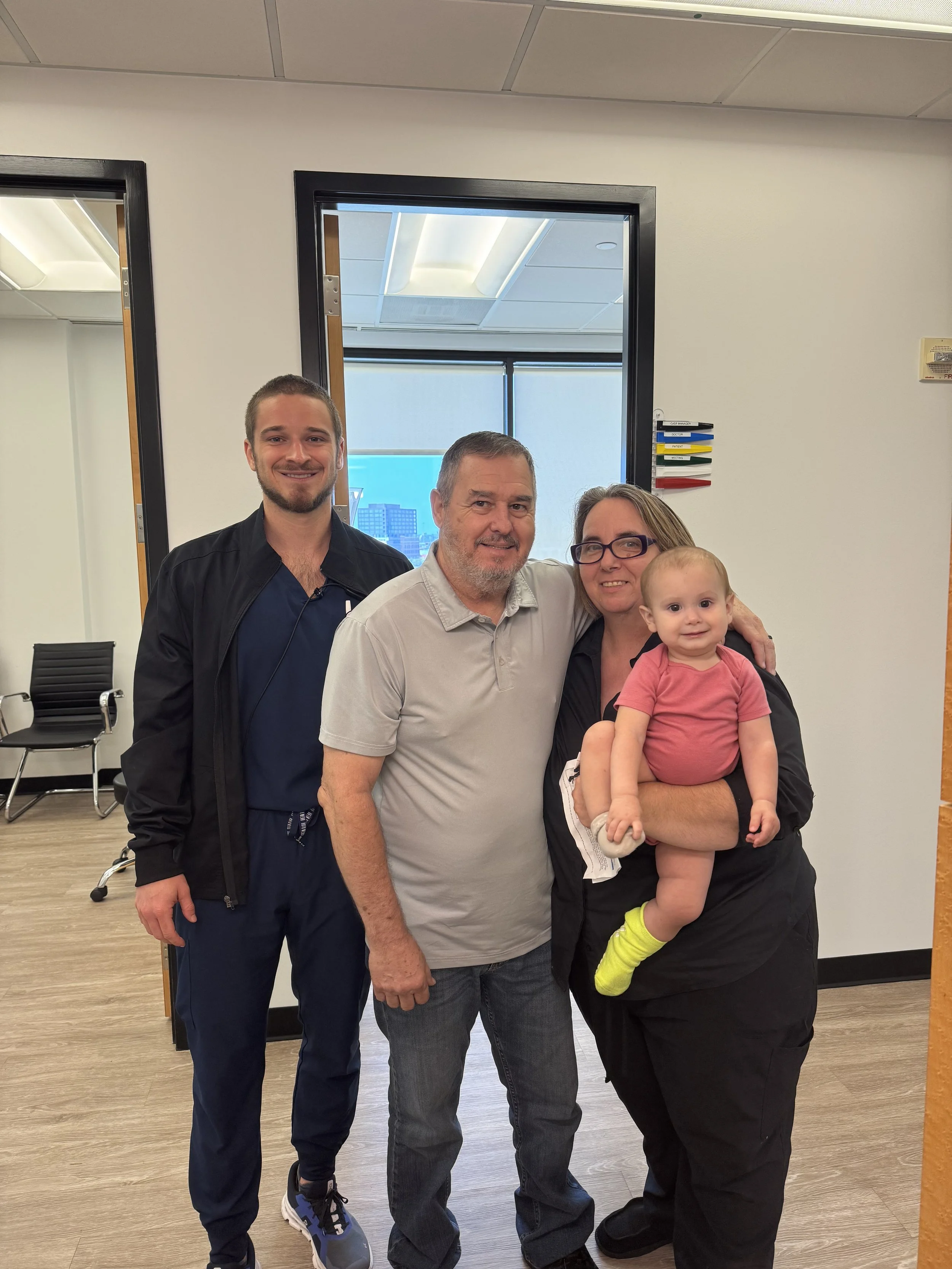 A family of four standing in a room, smiling at the camera. The father, mother, and a young man are in the photo, with the mother holding a toddler girl who is sitting on her arm.