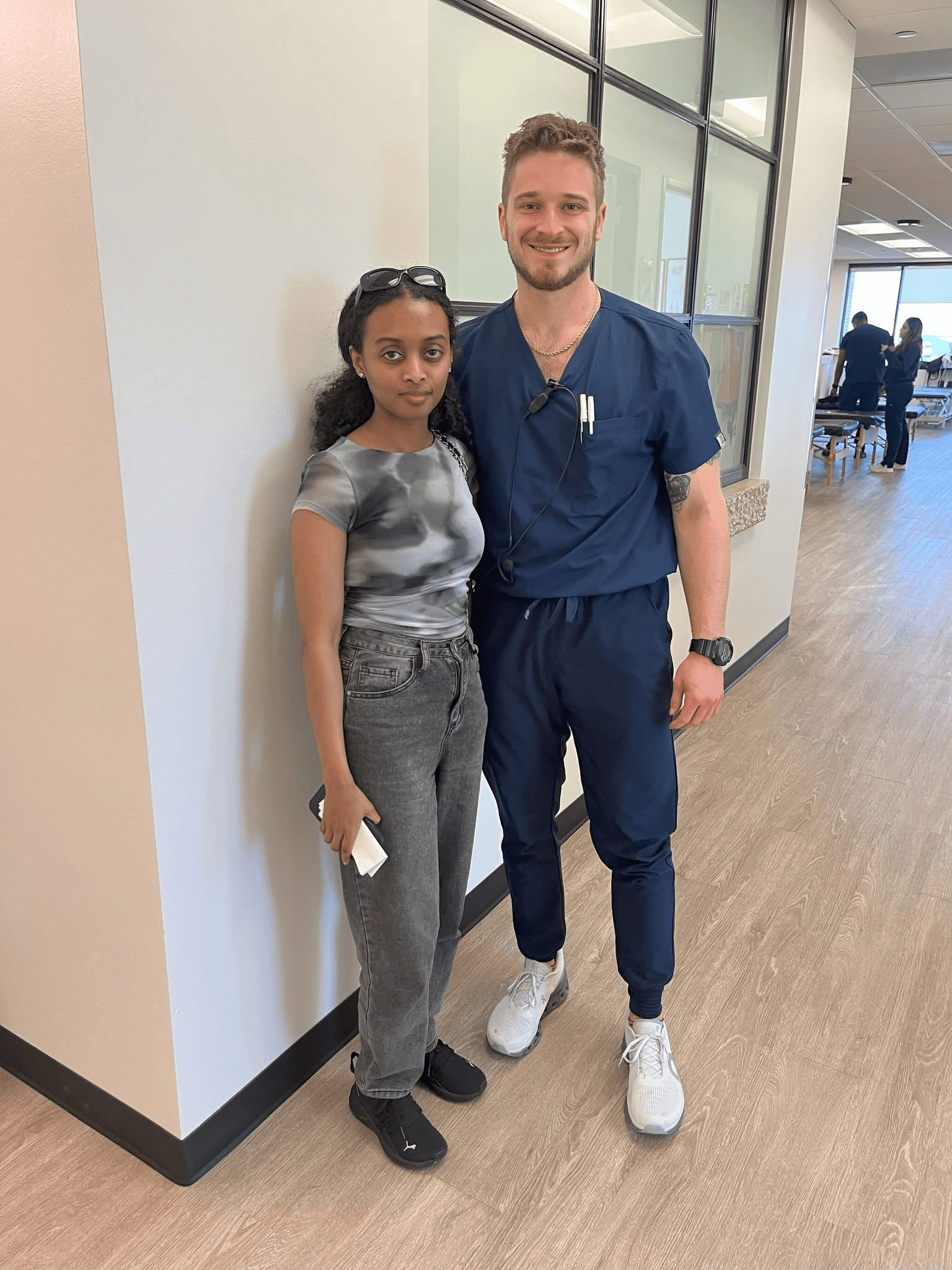 A woman and a man, possibly a healthcare professional, standing together in a hospital corridor, both smiling at the camera.