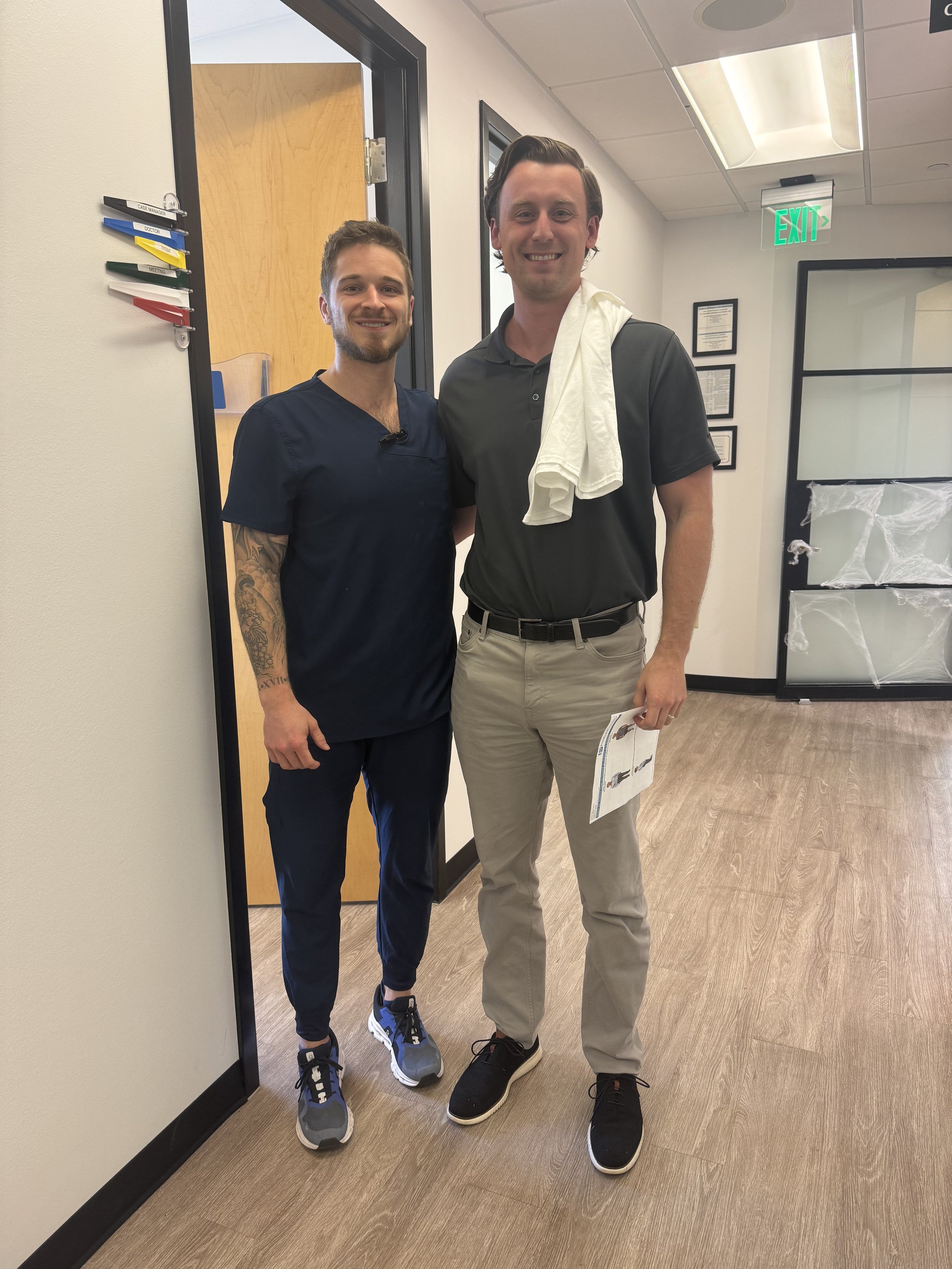 Two men standing together in an office, smiling at the camera. One is wearing medical scrubs, the other casual clothing with a white towel over his shoulder, holding a paper.