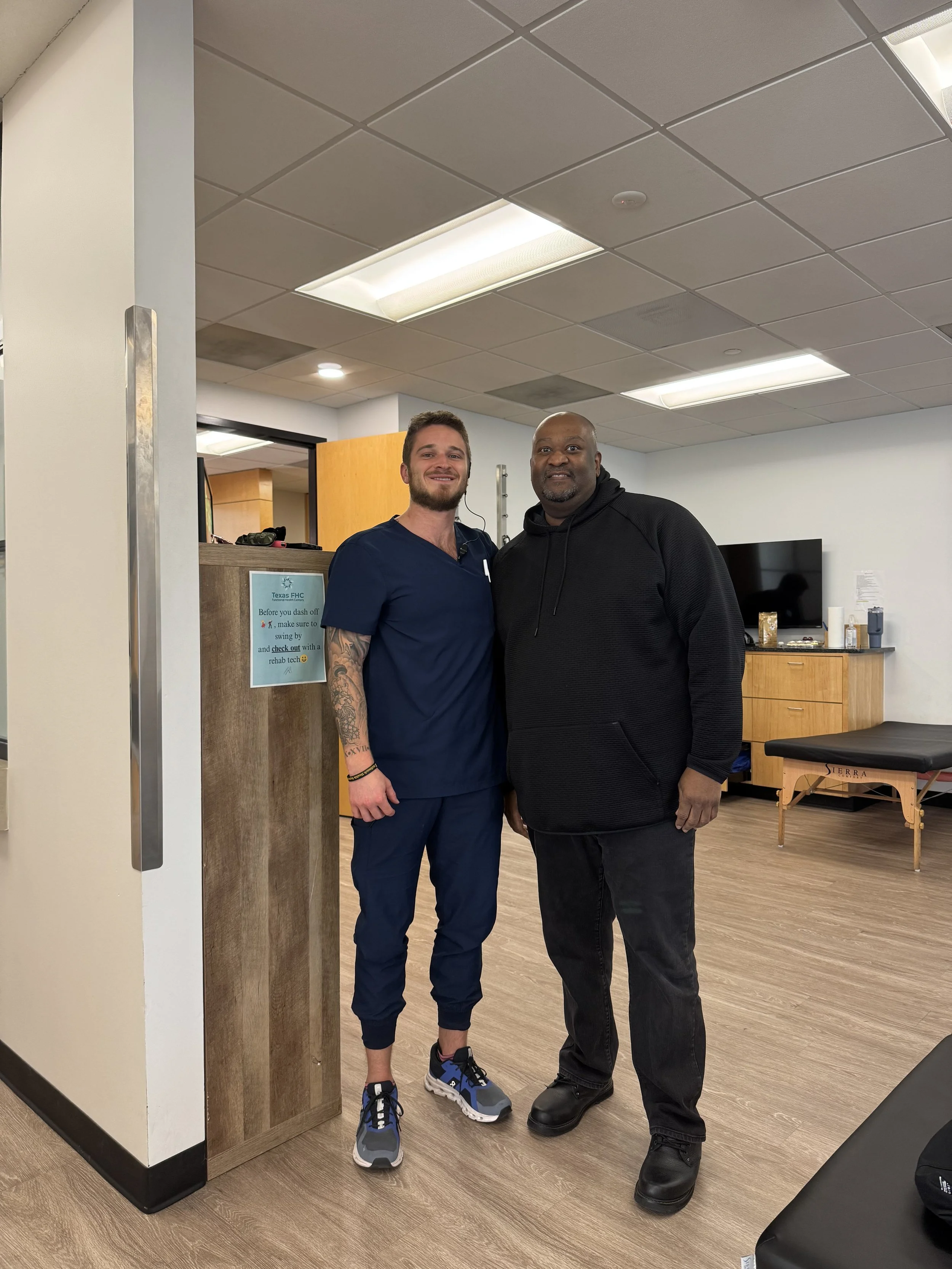 Two men, one in medical scrubs and the other in a black hoodie, standing together inside a medical office or clinic, smiling at the camera.