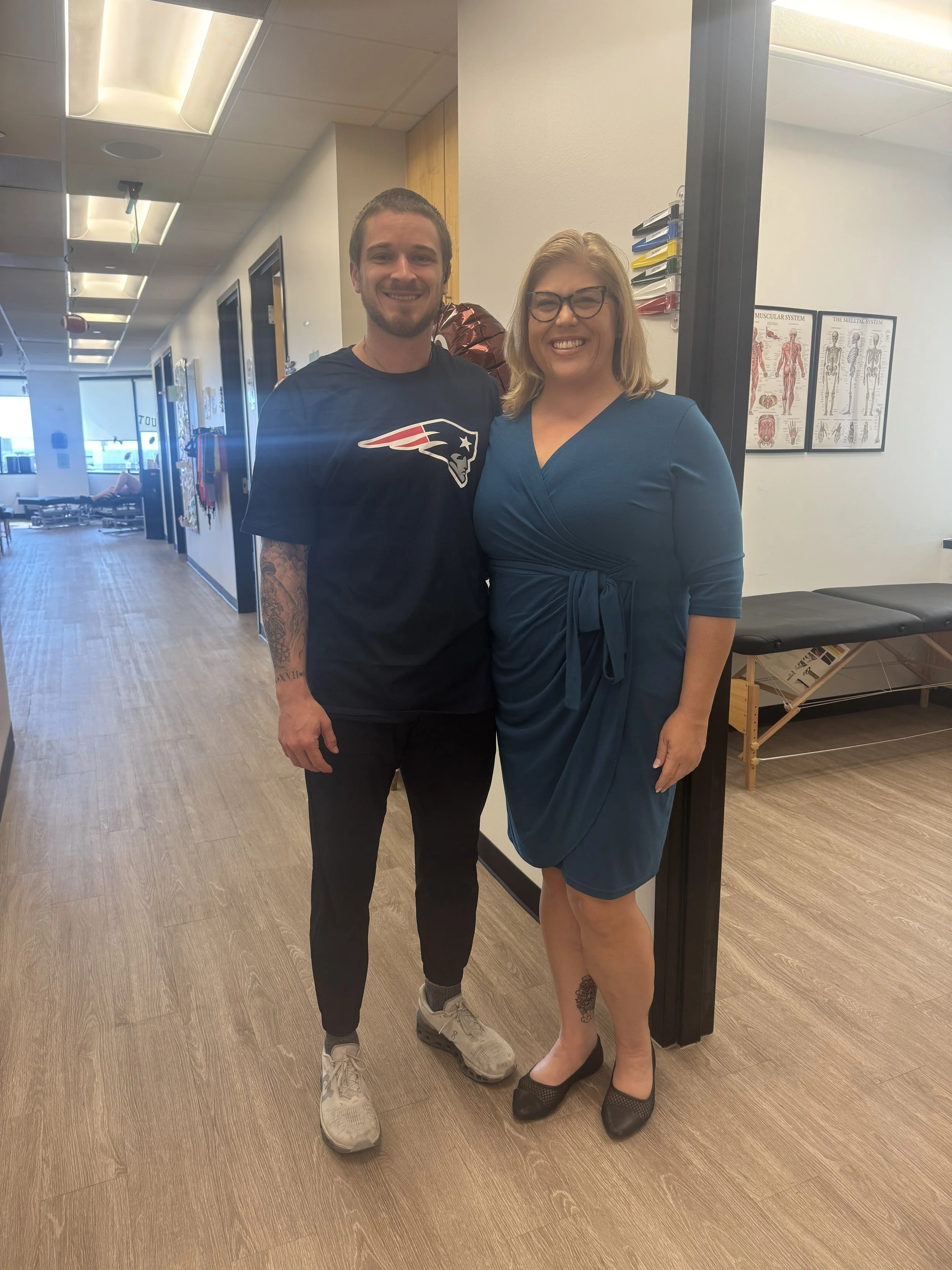 A young man and a woman standing together in a healthcare or wellness facility. The young man is wearing a New England Patriots t-shirt, black pants, and sneakers, with visible tattoo on his left arm. The woman, dressed in a teal wrap dress with a tattoo on her left ankle, is smiling. They are standing close to each other in front of a doorway with medical charts on the wall behind them.