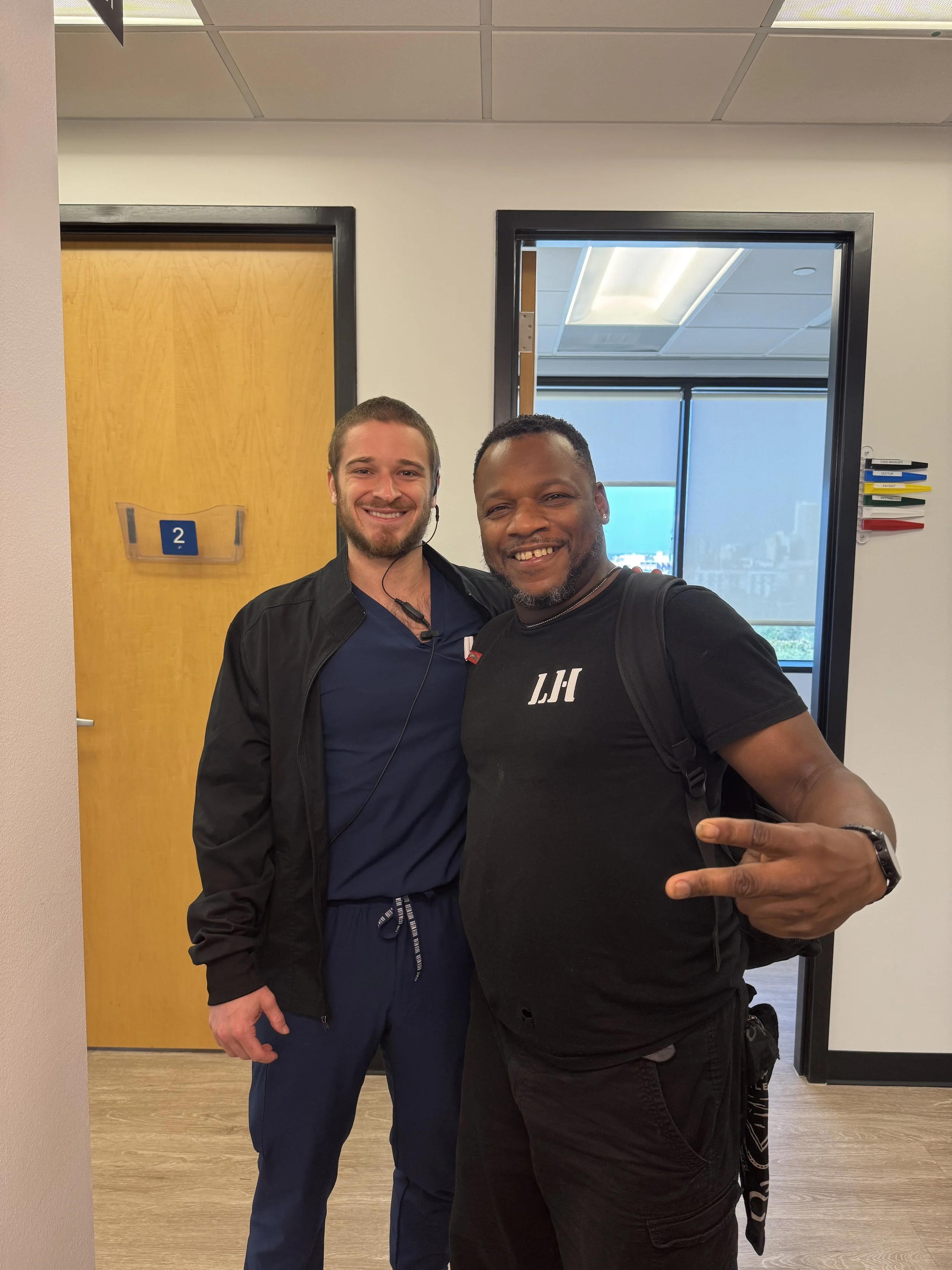 Two smiling men posing together indoors, one wearing scrubs and a black jacket, the other in a black t-shirt with 'LH' and a backpack. They are standing in front of a wooden door marked with a blue number two, with a window and cityscape visible in the background.
