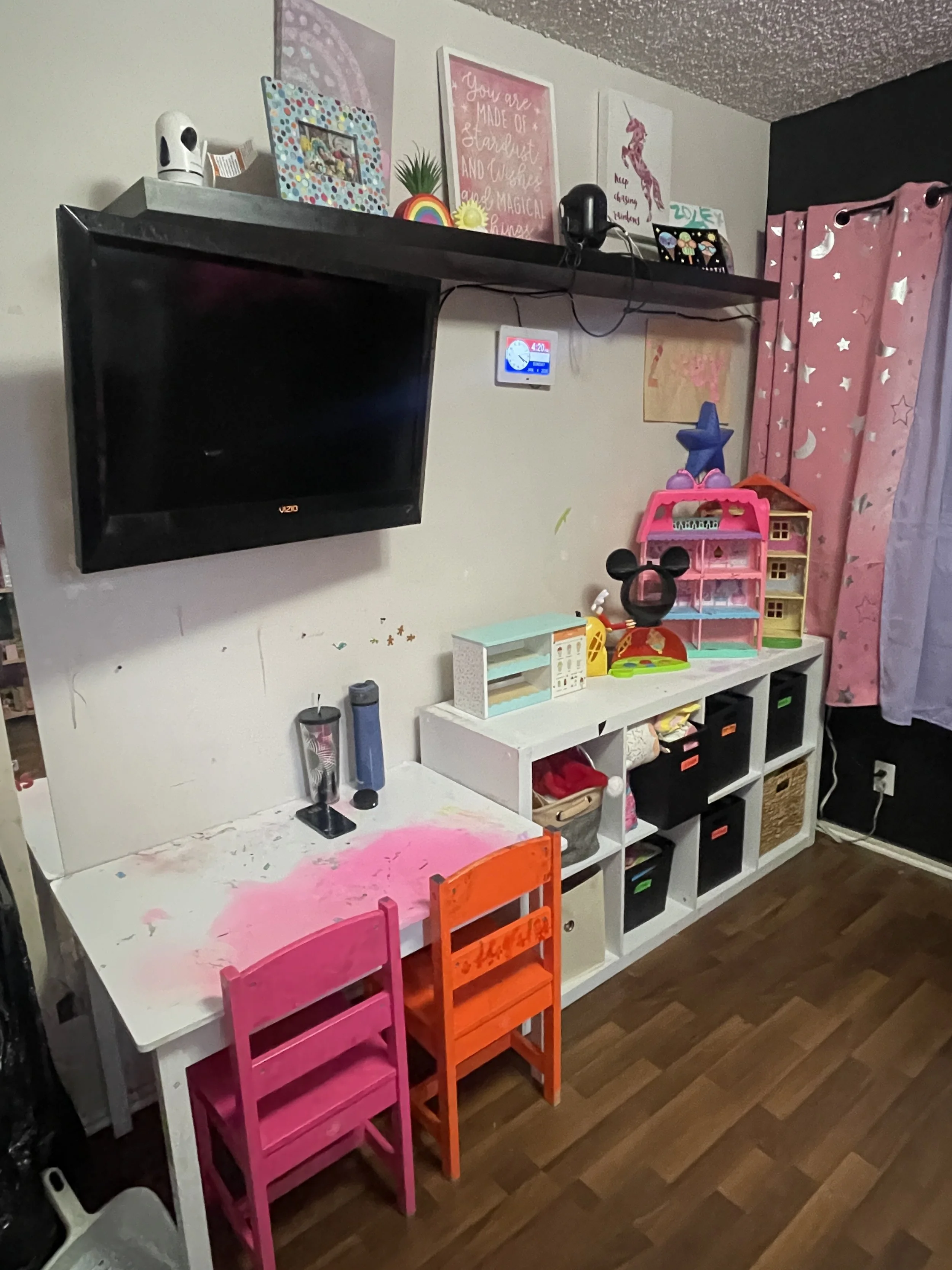 A children's playroom with a white table and two colorful chairs, a wall-mounted TV, a white cube shelving unit filled with toys and storage bins, pink curtains with star and moon patterns, and wall decorations including pictures and a motivational quote.