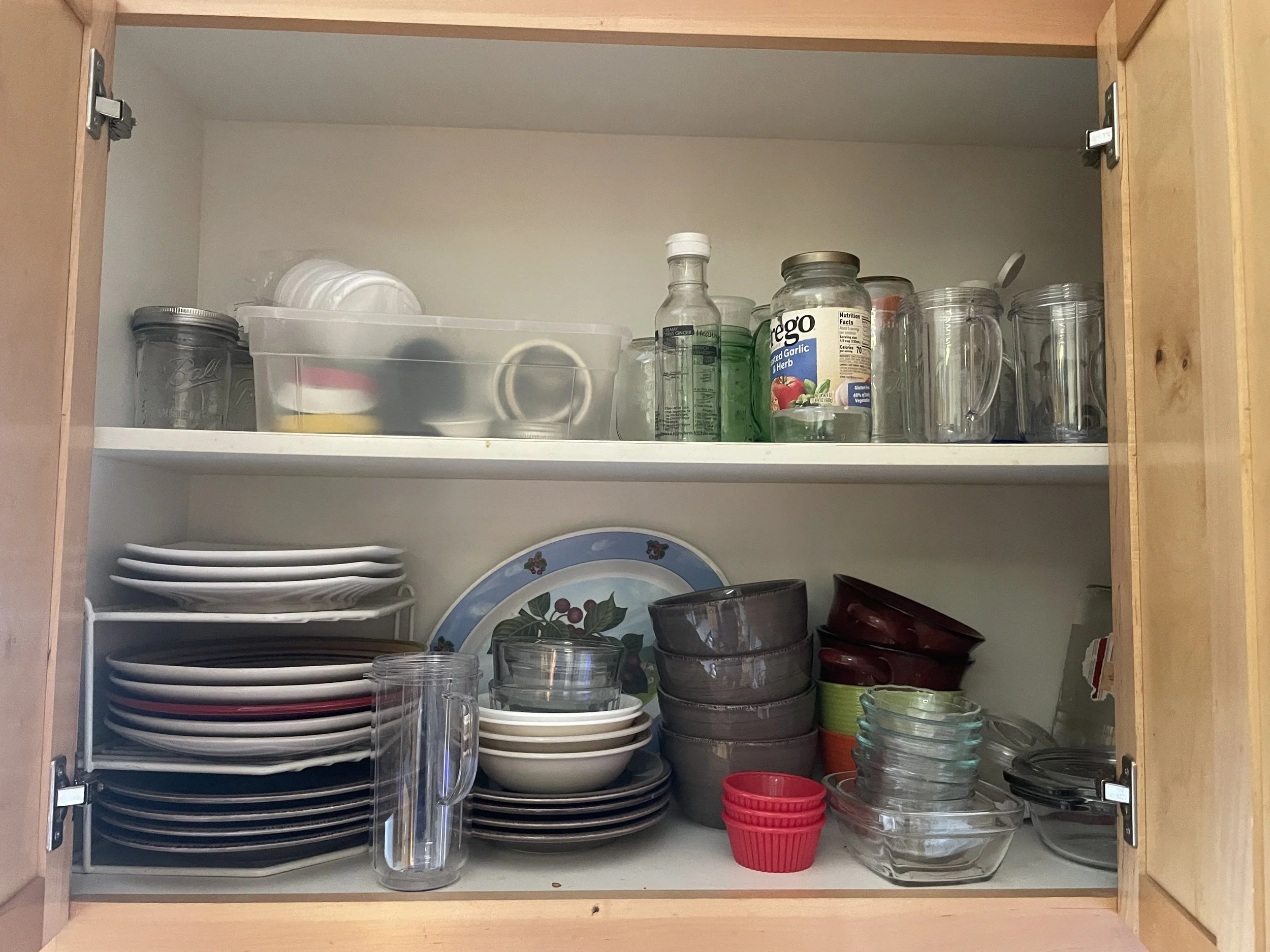 An open kitchen cabinet containing dishes, bowls, glasses, and food containers.
