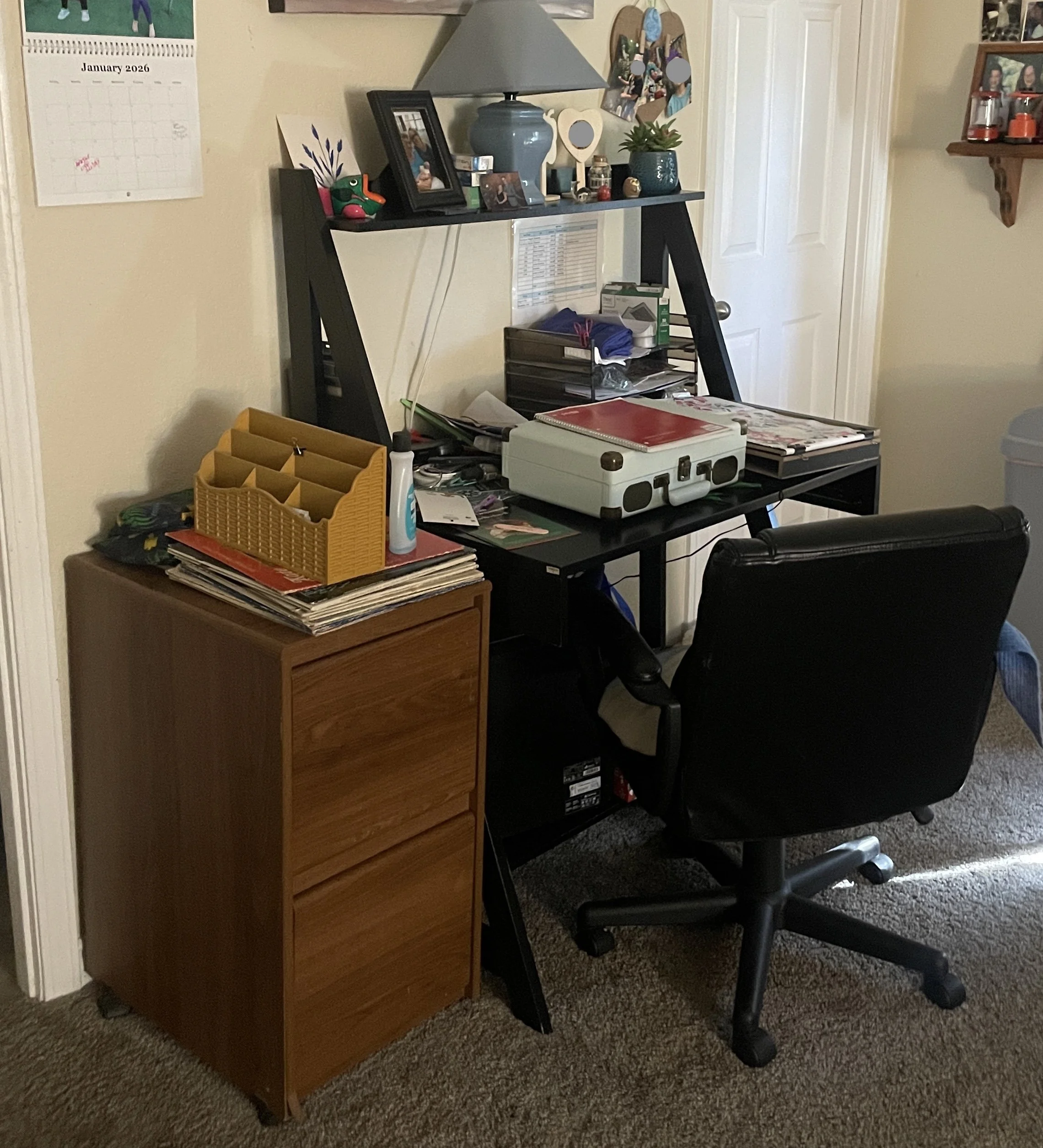 A workspace with a black desk and a wooden filing cabinet. The desk holds various items including a white briefcase, stacks of papers, a red notebook, and office supplies. There is a black office chair in front of the desk. Behind the desk, attached to the wall, is a black triangular shelving unit with personal items, framed photos, a blue lamp, and small decorative objects. To the left, there is a wall calendar for January 2026, and to the right, a closed white closet door and a wooden wall shelf with jars and decorations.