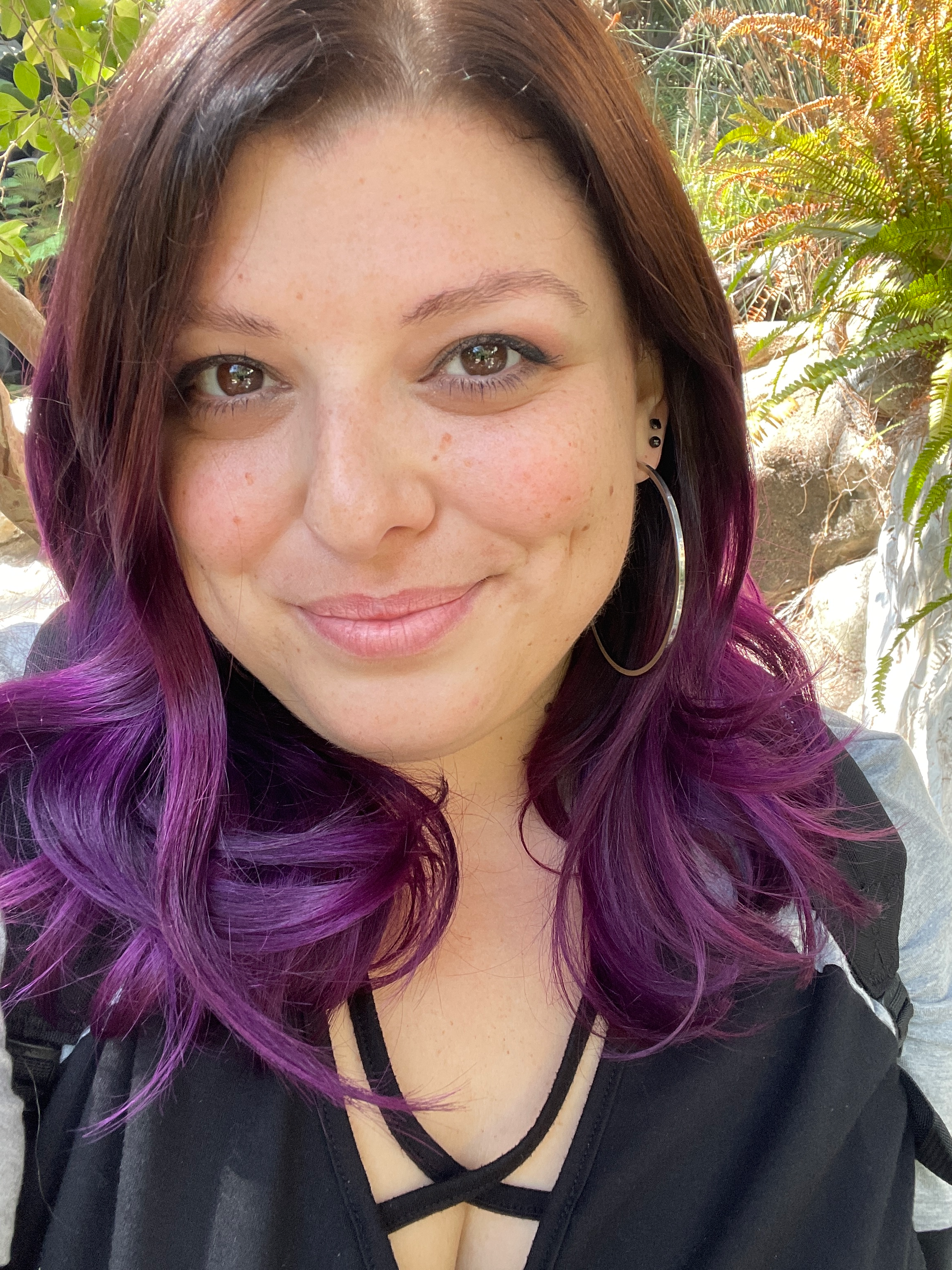 A young woman with brown and purple wavy hair, wearing hoop earrings, smiling outdoors with green plants in the background.