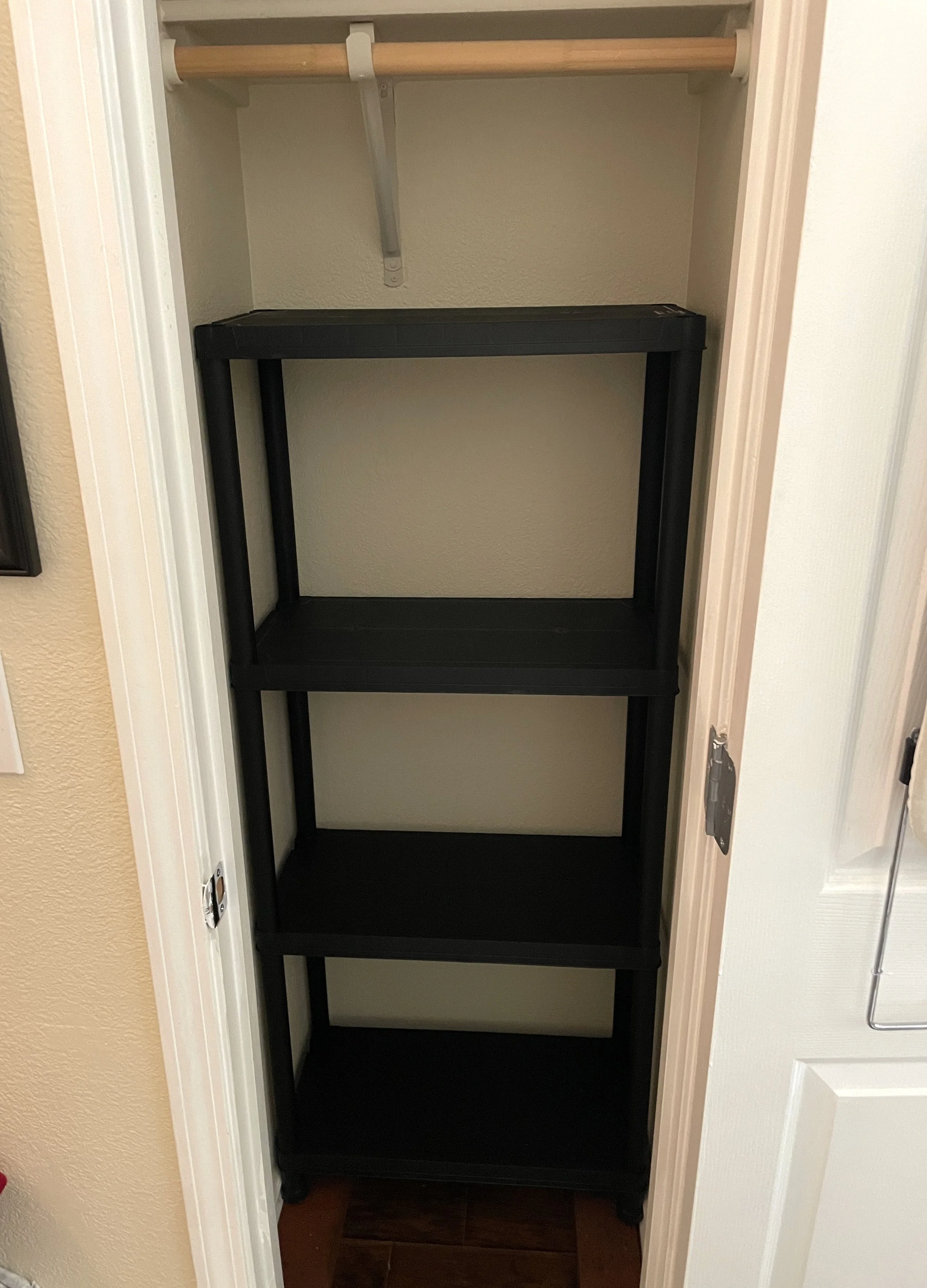 A small, empty black shelving unit with four shelves, placed inside a closet with a wooden rod and an adjustable white metal support, with beige walls and brown tile flooring.