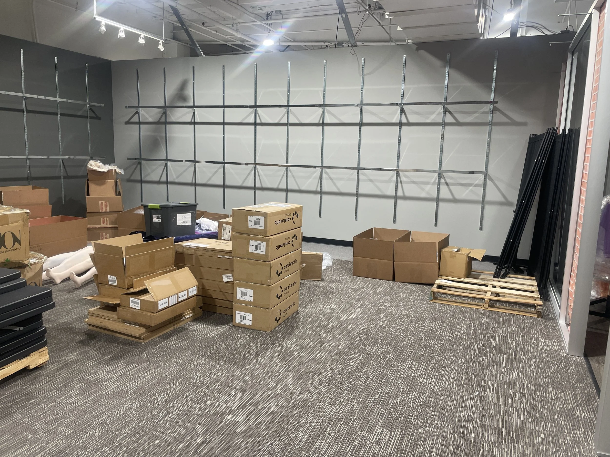 Empty retail store or display area with metal wall fixtures and shelves, stacked cardboard boxes, some pallets, and black metal rods on the floor.
