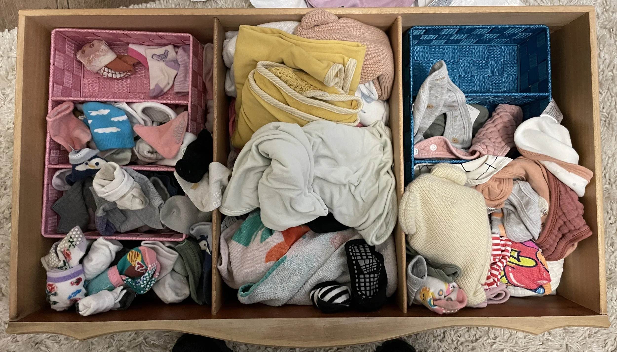 An open wooden drawer divided into three sections containing assorted baby socks, hats, and clothing, with colorful socks and neatly folded garments.