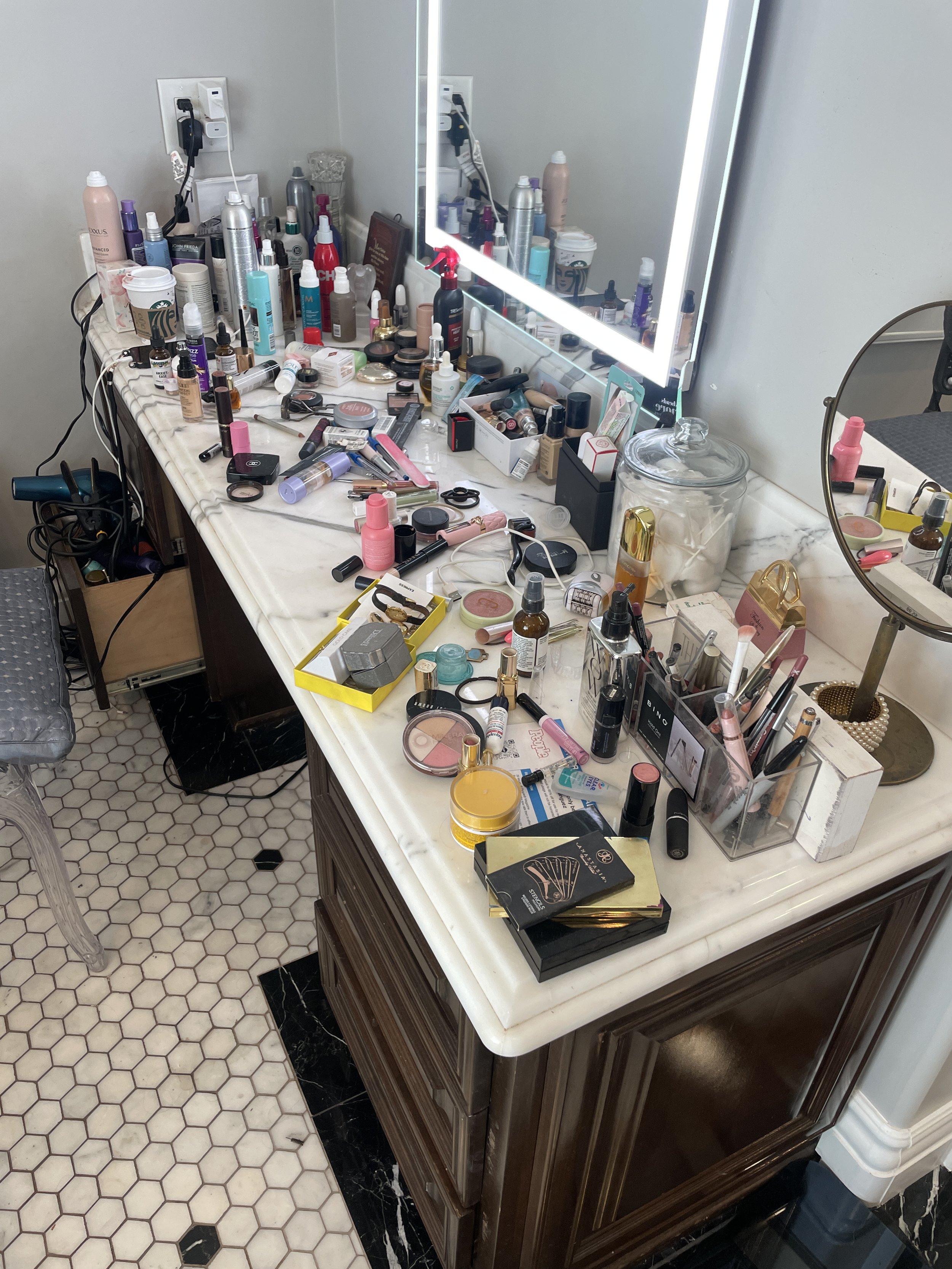A marble countertop filled with numerous makeup products, brushes, and tools. The mirror is illuminated with bright lighting, and there's a small round mirror to the right. The counter sits on a dark wood cabinet, with a tiled hexagon floor beneath.