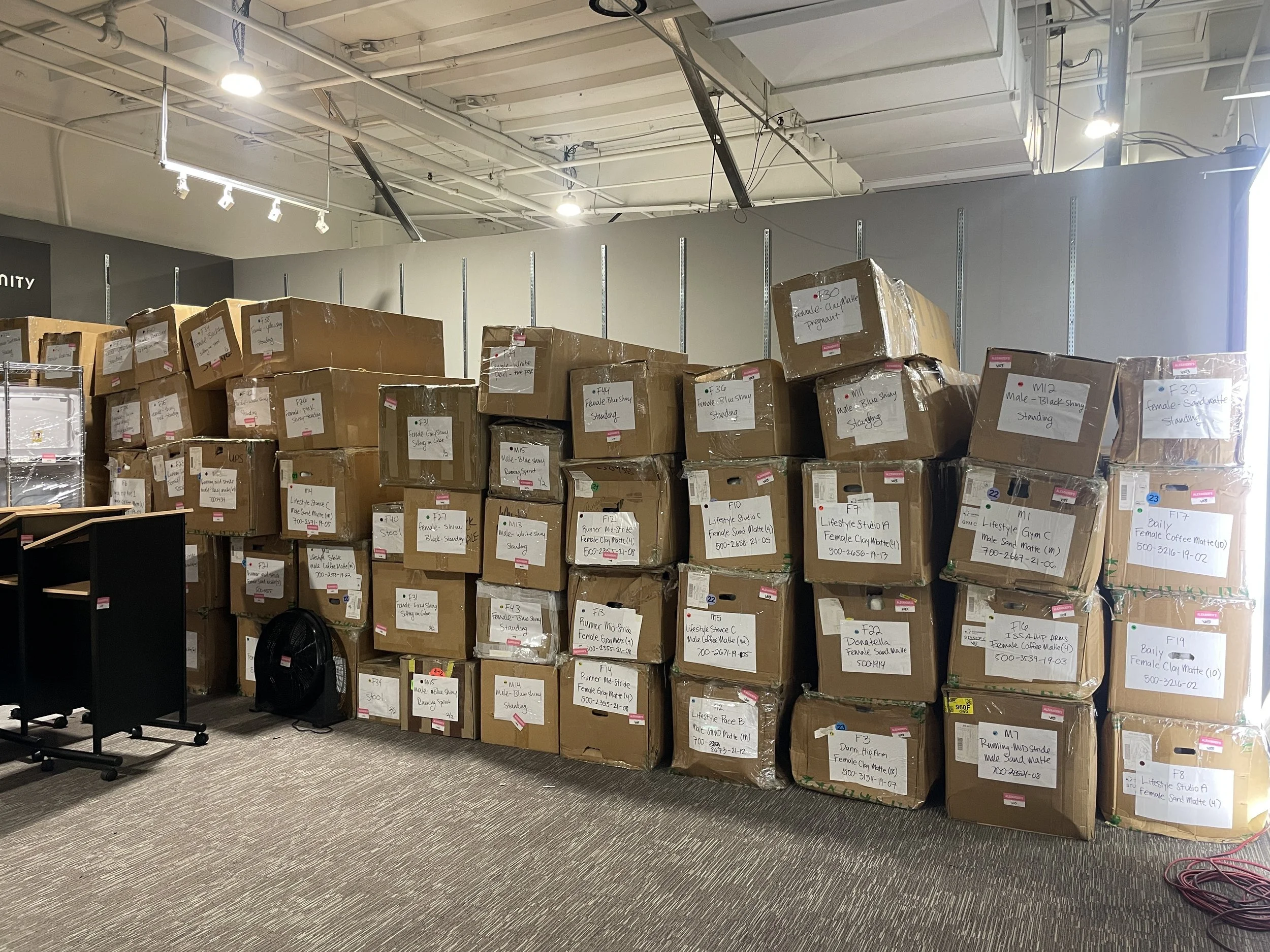 Large stacks of labeled cardboard boxes stored against a wall in an indoor space, with some furniture and a floor fan visible in the foreground.