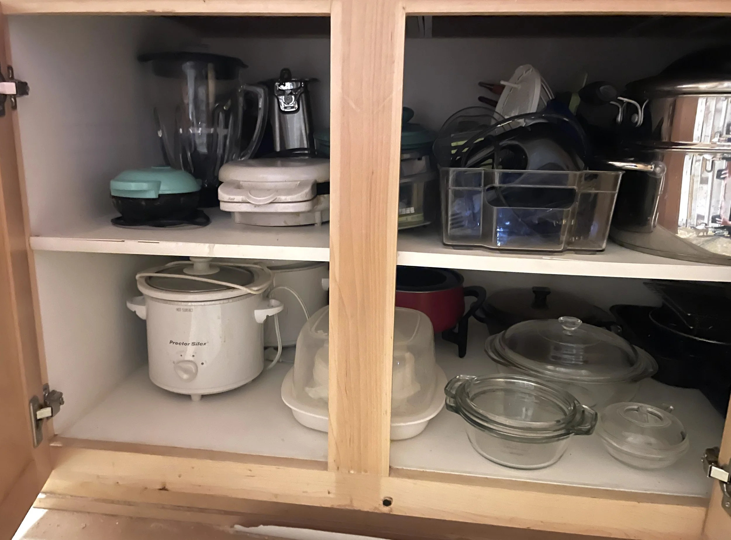 Kitchen cabinet filled with cookware, glassware, and small appliances. Left side has stacked bowls, a blender, and a rice cooker. Right side has pots, pans, and glass baking dishes.
