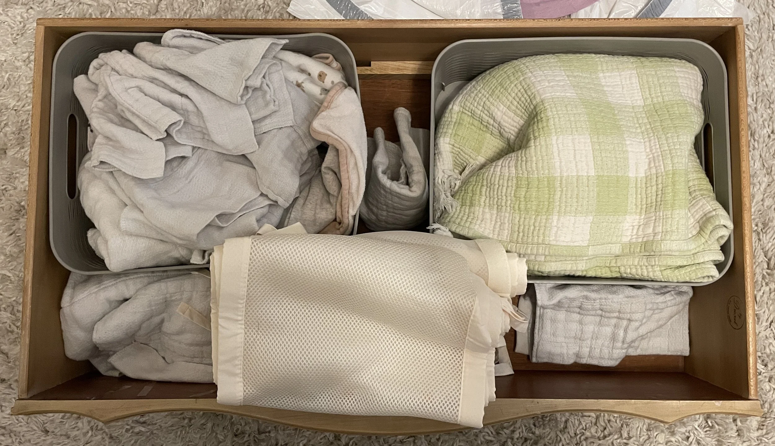 A wooden dresser drawer divided into sections containing folded white and light-colored baby clothes, a white mesh laundry bag, and a small green and white checked blanket.