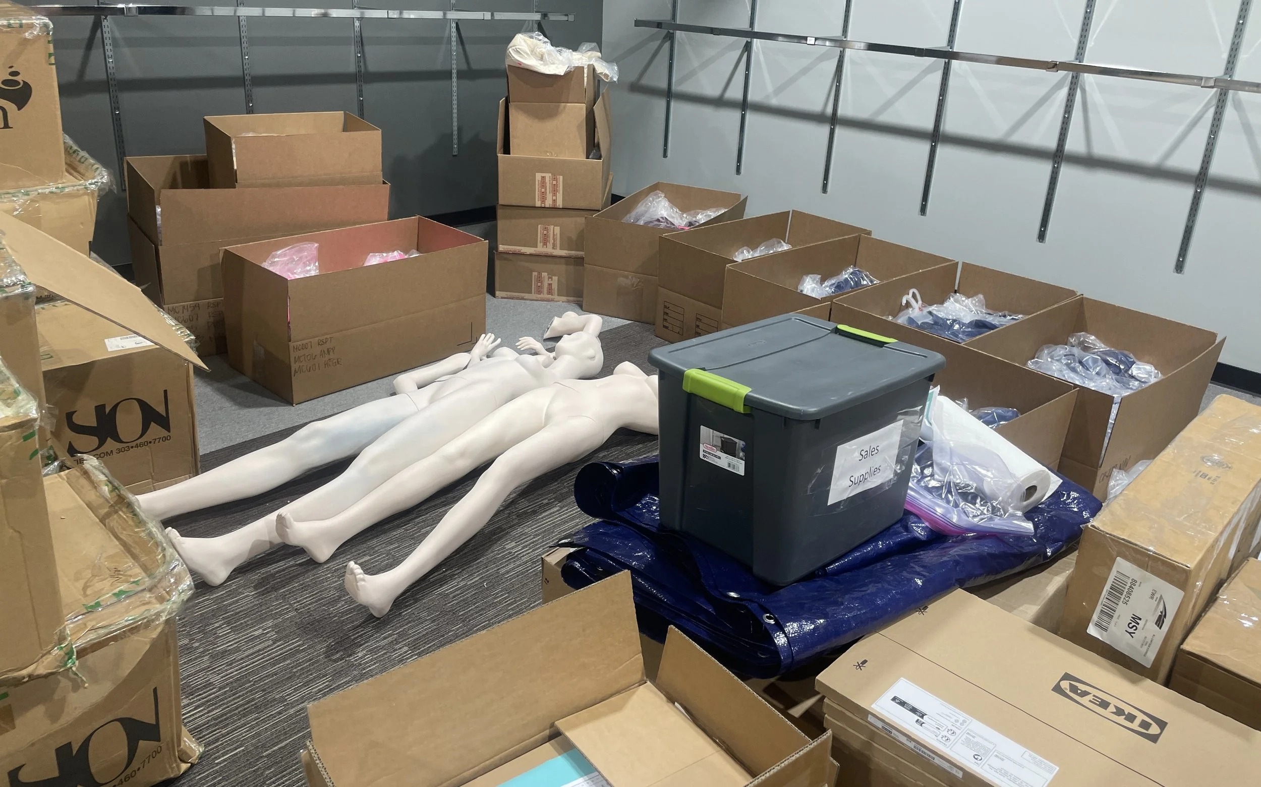 A storage room with several cardboard boxes, some open with bubble wrap, and three white mannequin torsos lying on the floor. There is a gray plastic container labeled 'Sales Supplies' and a rolled-up blue tarp on the floor.