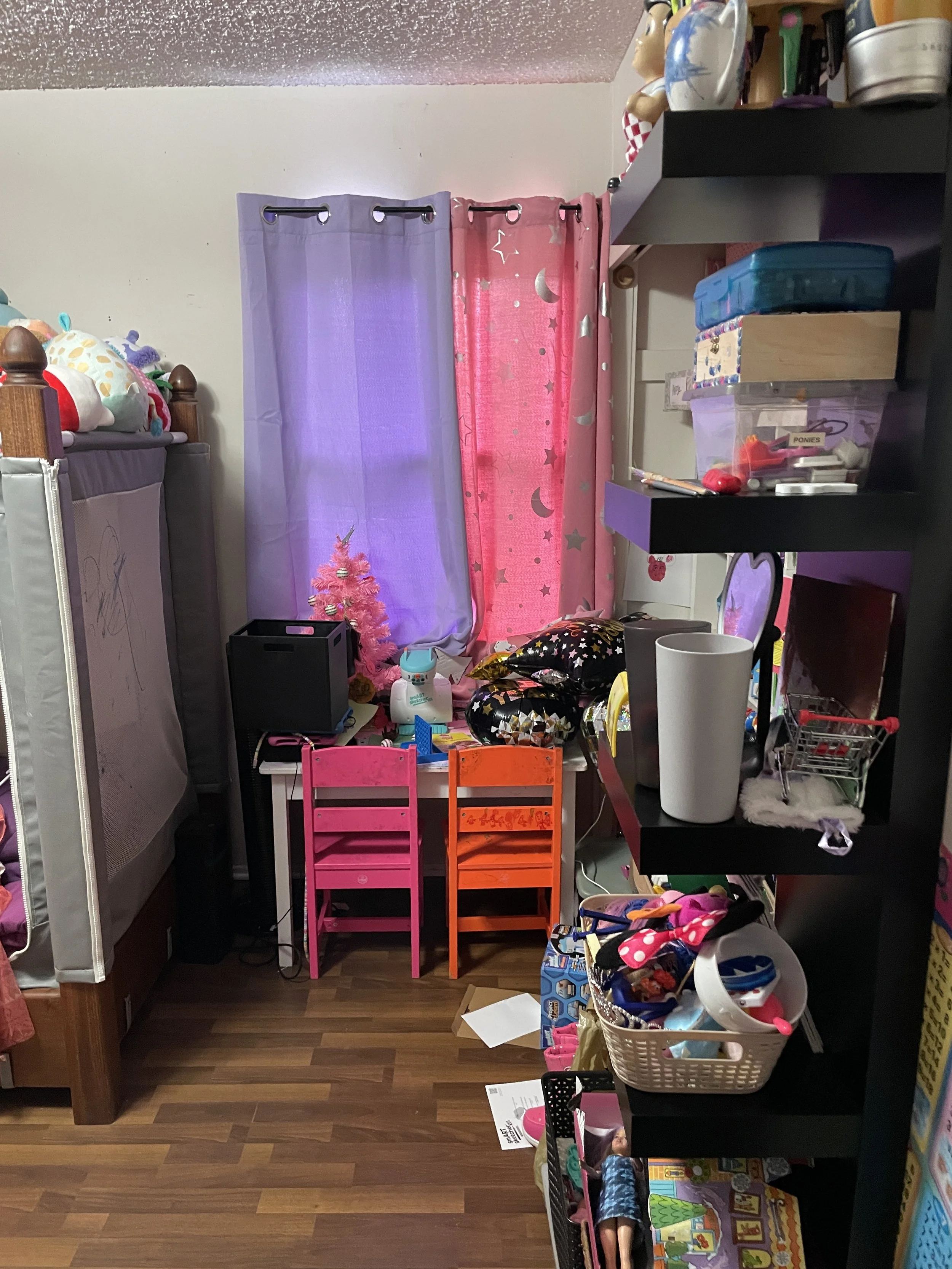 A cluttered corner of a child's room with colorful curtains, pink and orange chairs, a small pink Christmas tree, and various toys and storage bins.