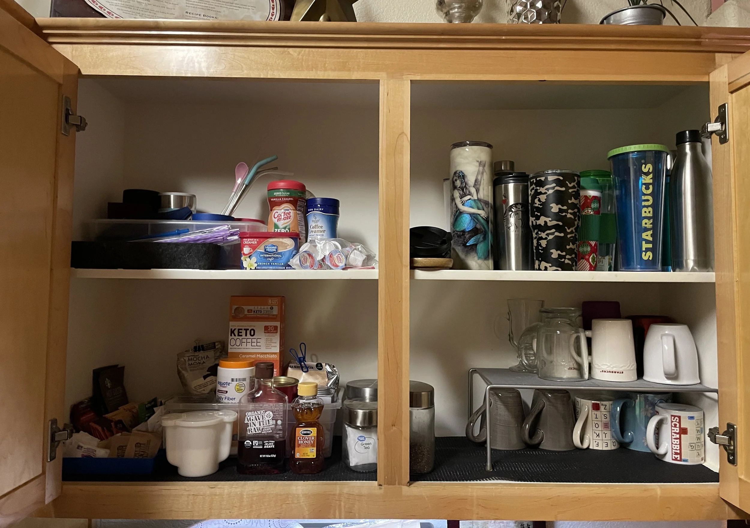 Open kitchen cabinet containing coffee, tea, mugs, and travel cups on the middle and bottom shelves, with various containers and kitchen accessories inside.